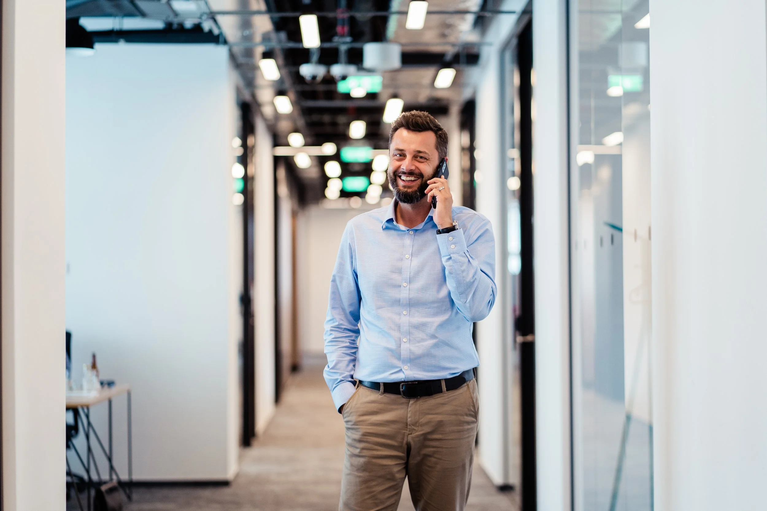 Smiling man in business casual attire talking on a mobile phone in a modern office corridor. Professional business headshot, personal branding, and corporate startup in Zürich, Zug, Bern, Lucerne, Basel, St. Gallen and across Switzerland.