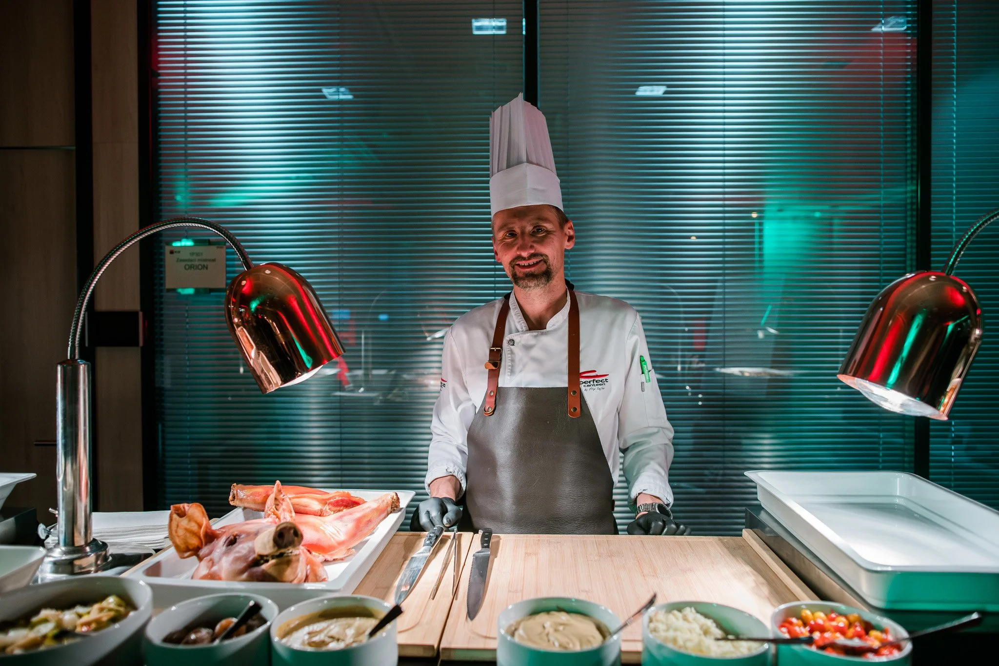 Chef standing behind a wooden counter with ingredients, including raw meat and prepared dishes, in a professional kitchen with modern background. Catering and food photography in Zürich, Zug, Bern, Lucerne, Basel, St. Gallen and across Switzerland.