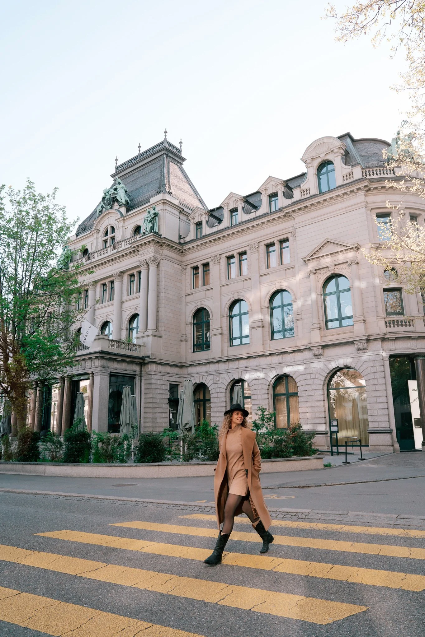 A woman crossing a street in front of a historic European-style building in St. Gallen, Switzerland, with ornate architectural details, surrounded by trees and greenery.