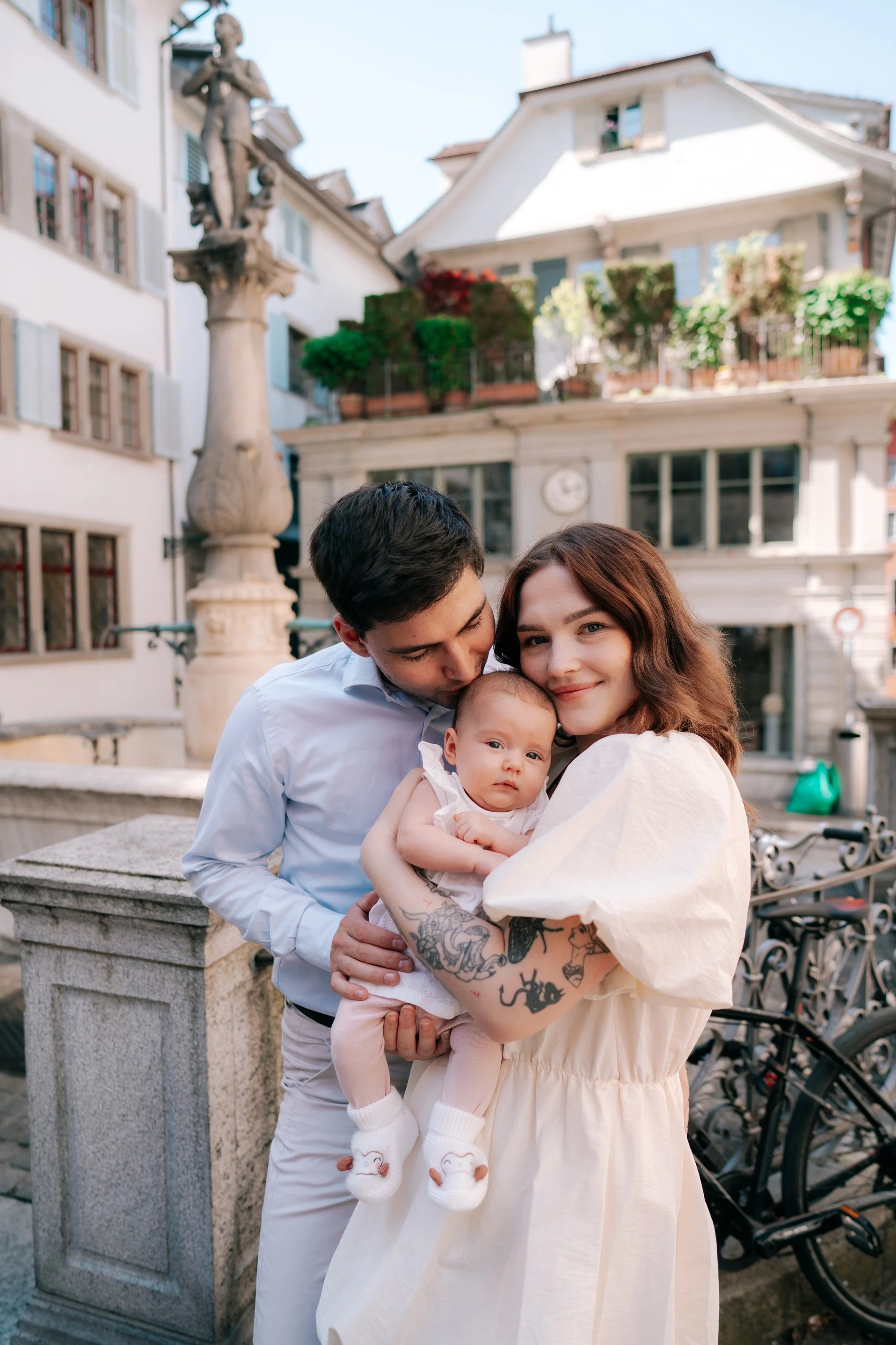 Young family with a baby outdoors lovingly taking photos in Zurich Switzerland, with historic buildings and a fountain in the background.