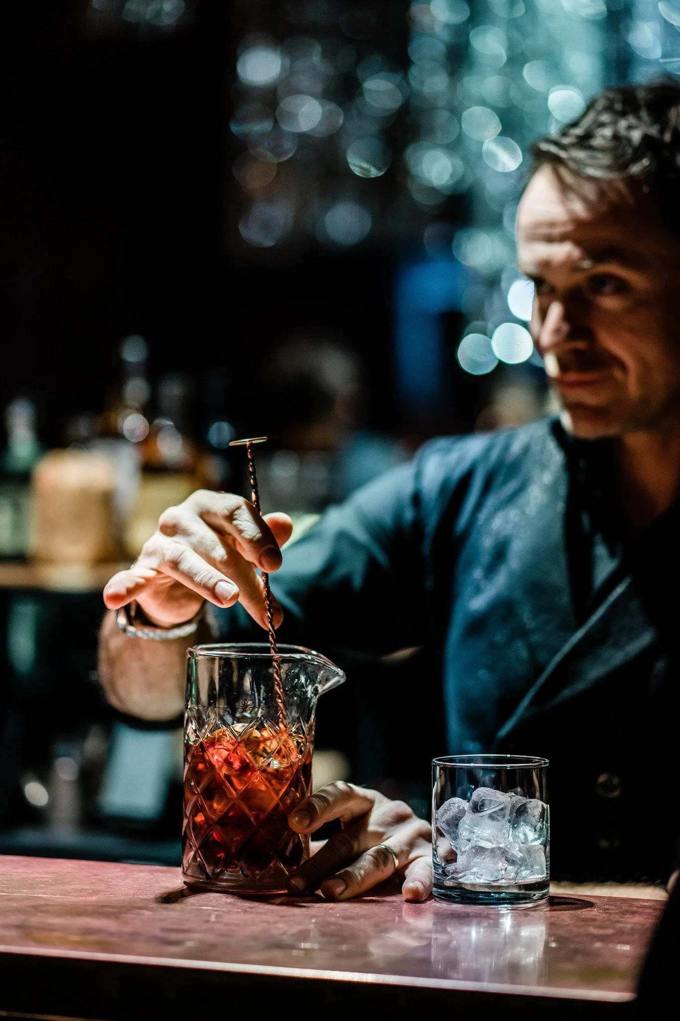 A bartender preparing a cocktail with a long spoon in a dimly lit bar, with a glass of ice and a pitcher on the wooden counter. Catering, culinary and food photography in Zürich, Zug, Bern, Lucerne, Basel, St. Gallen and across Switzerland.
