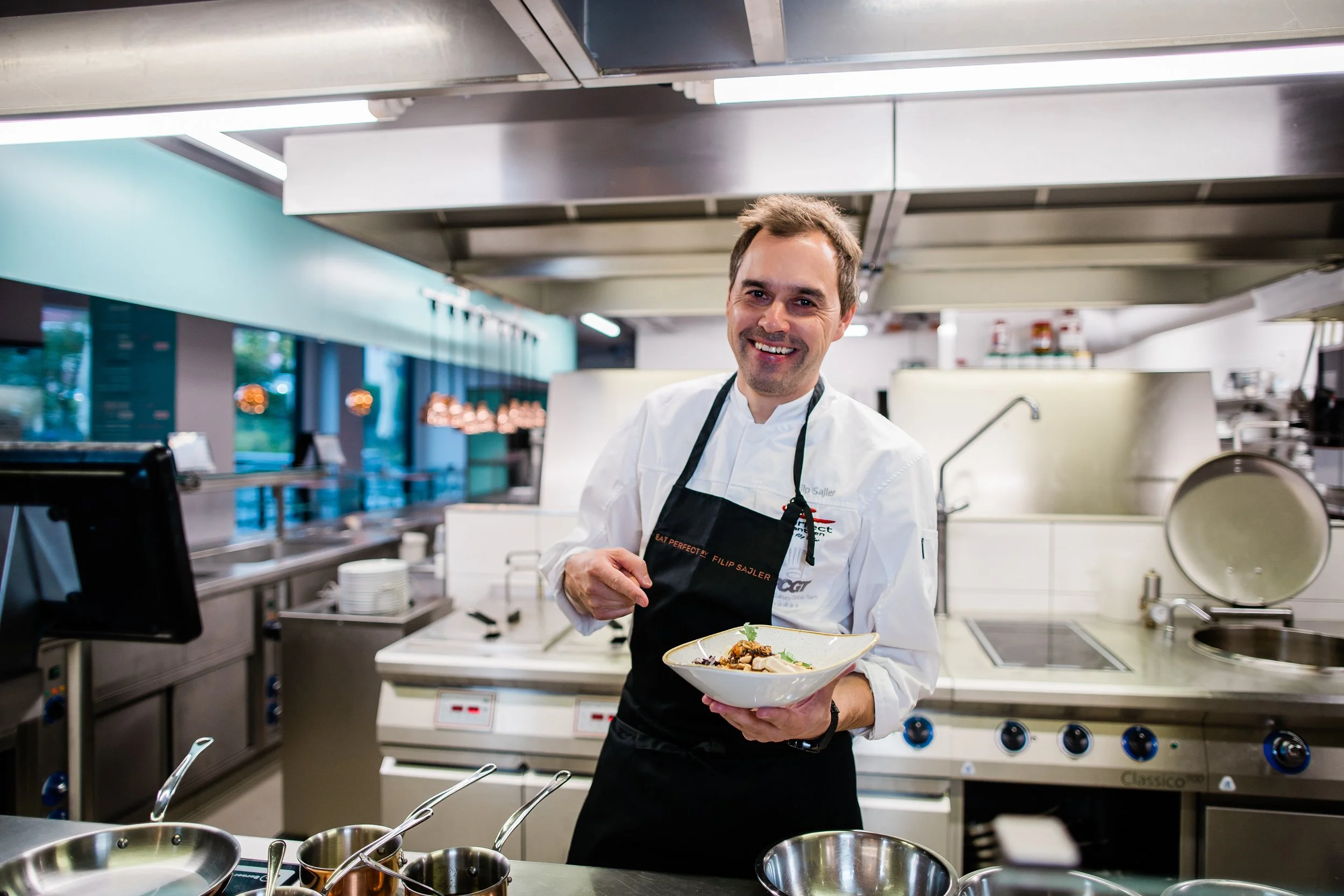 A smiling male chef in a white uniform and black apron holding a plated dish in a professional kitchen. Personal branding and fine dining restaurant photography in Zürich, Zug, Bern, Lucerne, Basel, St. Gallen and across Switzerland.
