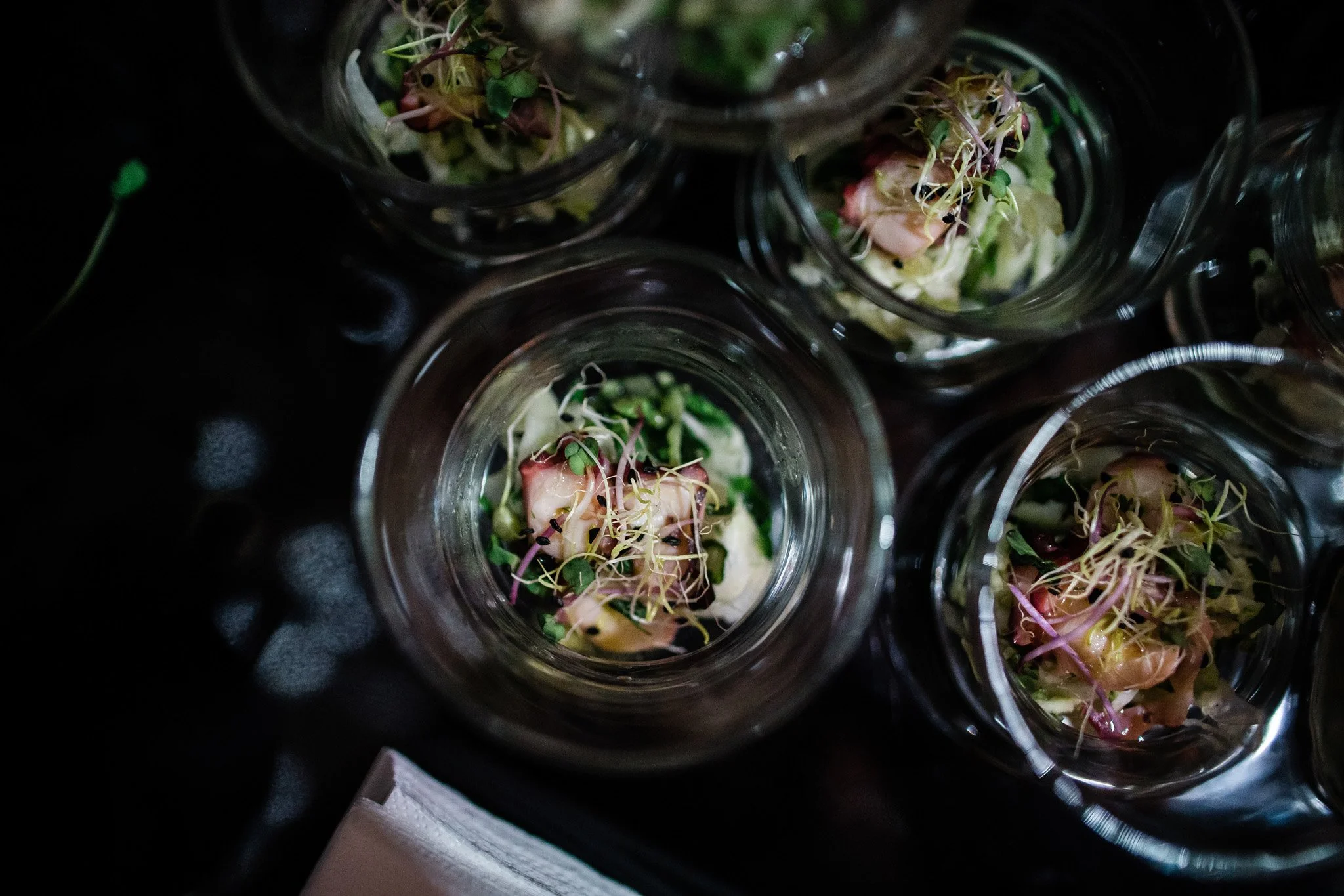 Multiple small glass jars containing a salad with greens, microgreens, and possibly avocado or egg, viewed from above on a dark surface. Catering and food photography in Zürich, Zug, Bern, Lucerne, Basel, St. Gallen and across Switzerland.