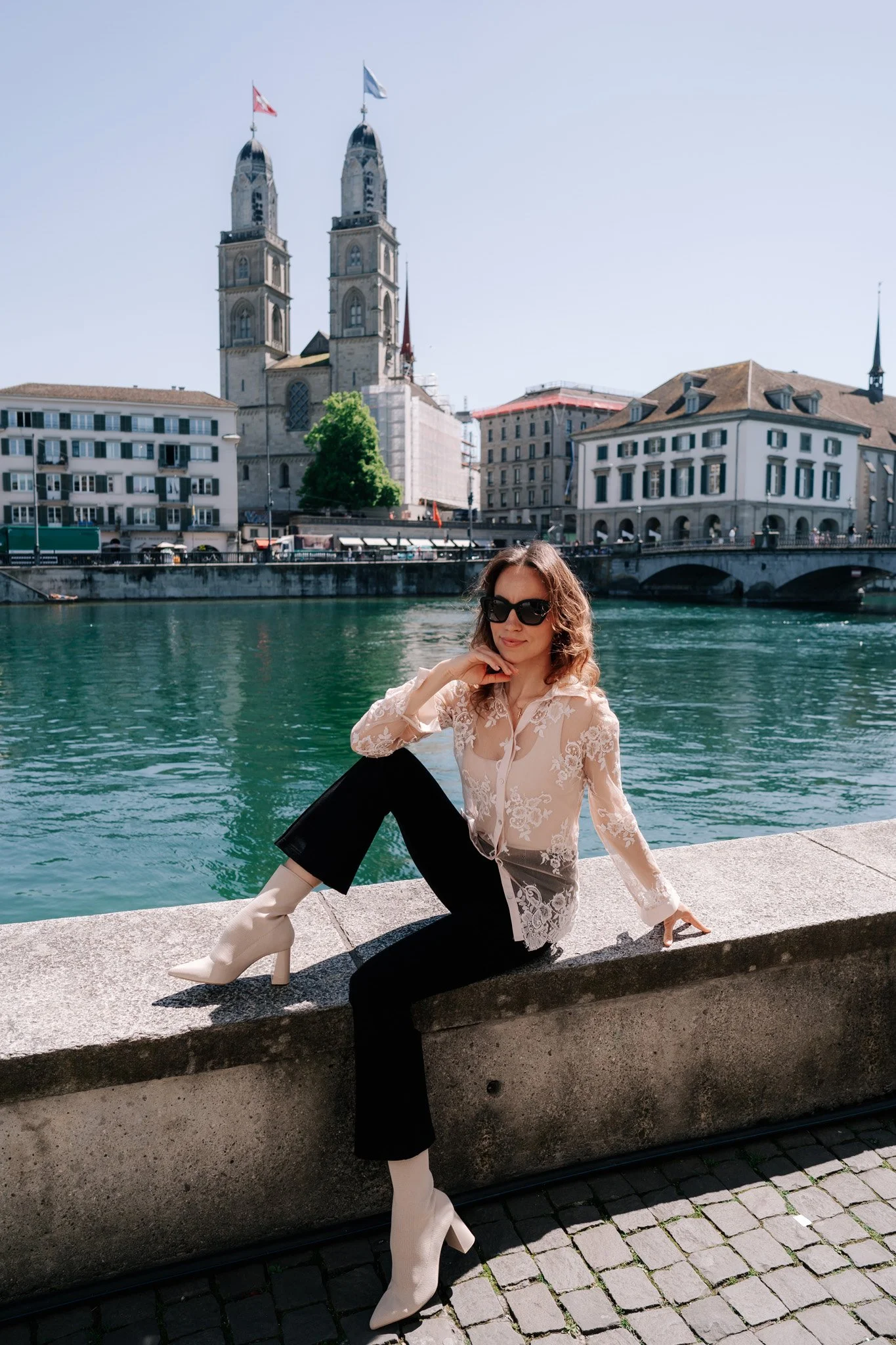 A woman taking solo travel photoshoot with curly hair and sunglasses sitting by a river in Zurich, with historic buildings and a church with twin towers in the background.
