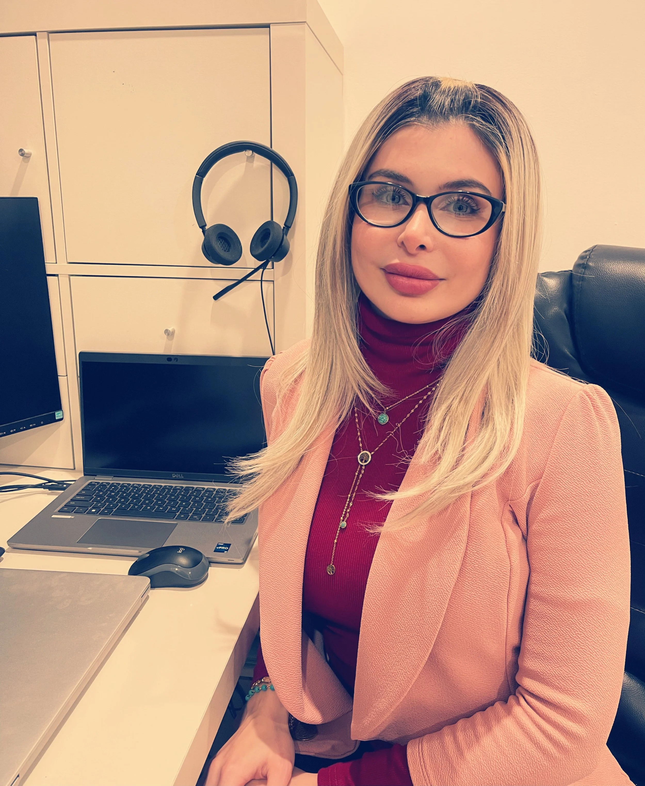 A woman with blonde hair and glasses sitting at a desk with a laptop, headphones hanging on a cabinet, and two monitors.