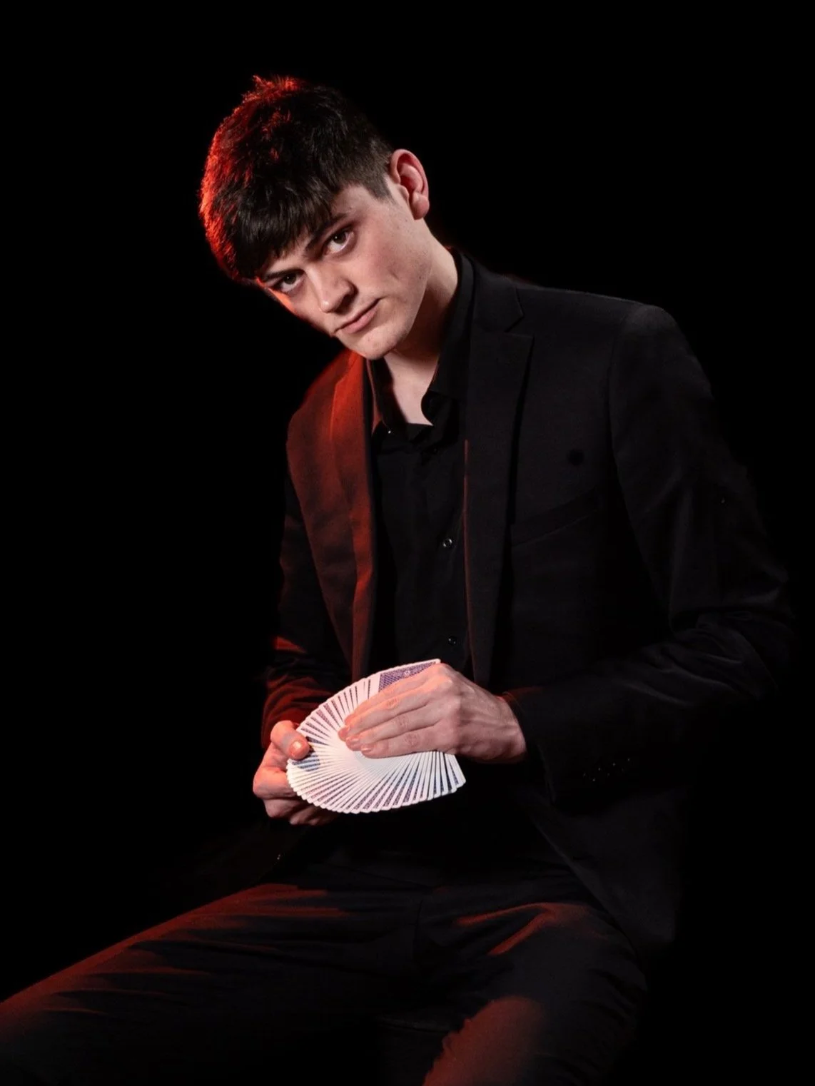 Young man dressed in black suit holding a fan of playing cards, sitting against a dark background with red lighting