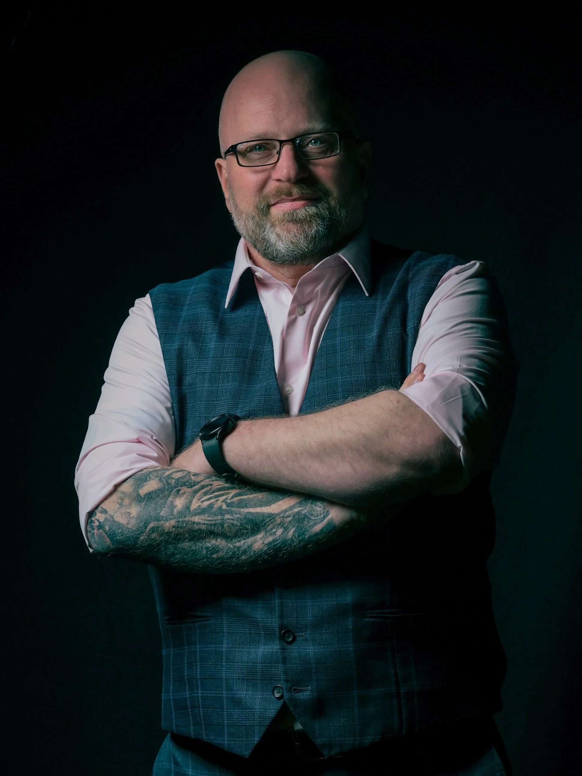 A middle-aged man with glasses, a beard, and tattoos on his left arm, standing with arms crossed against a dark background, wearing a pink shirt and a blue plaid vest.