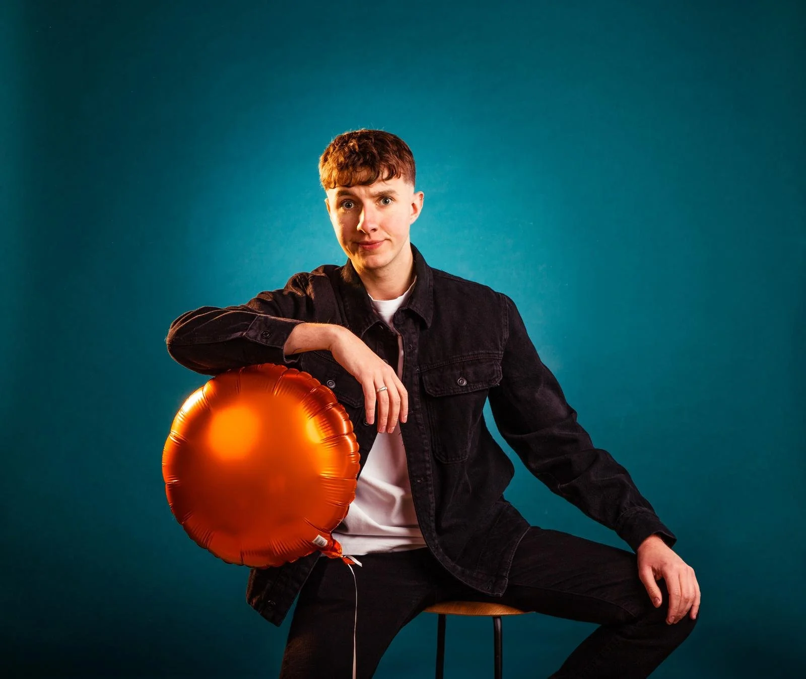 Young man with short red hair wearing a black denim jacket and white shirt, sitting on a stool with a mustard seat against a teal background, holding an orange foil balloon resembling a heart.