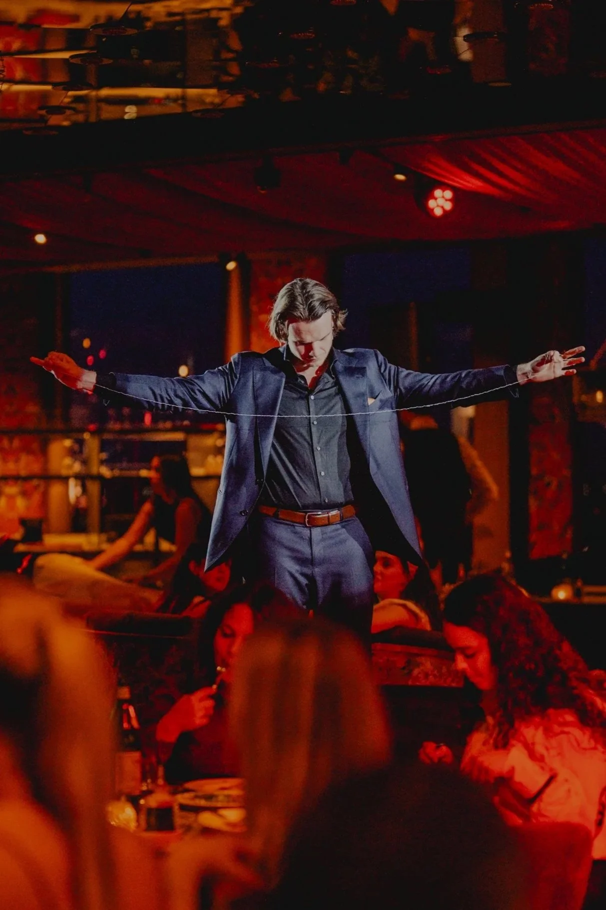 A man in a dark suit standing with arms outstretched on a stage, surrounded by a dimly lit crowd, in a vibrant lounge or club setting with red lighting.