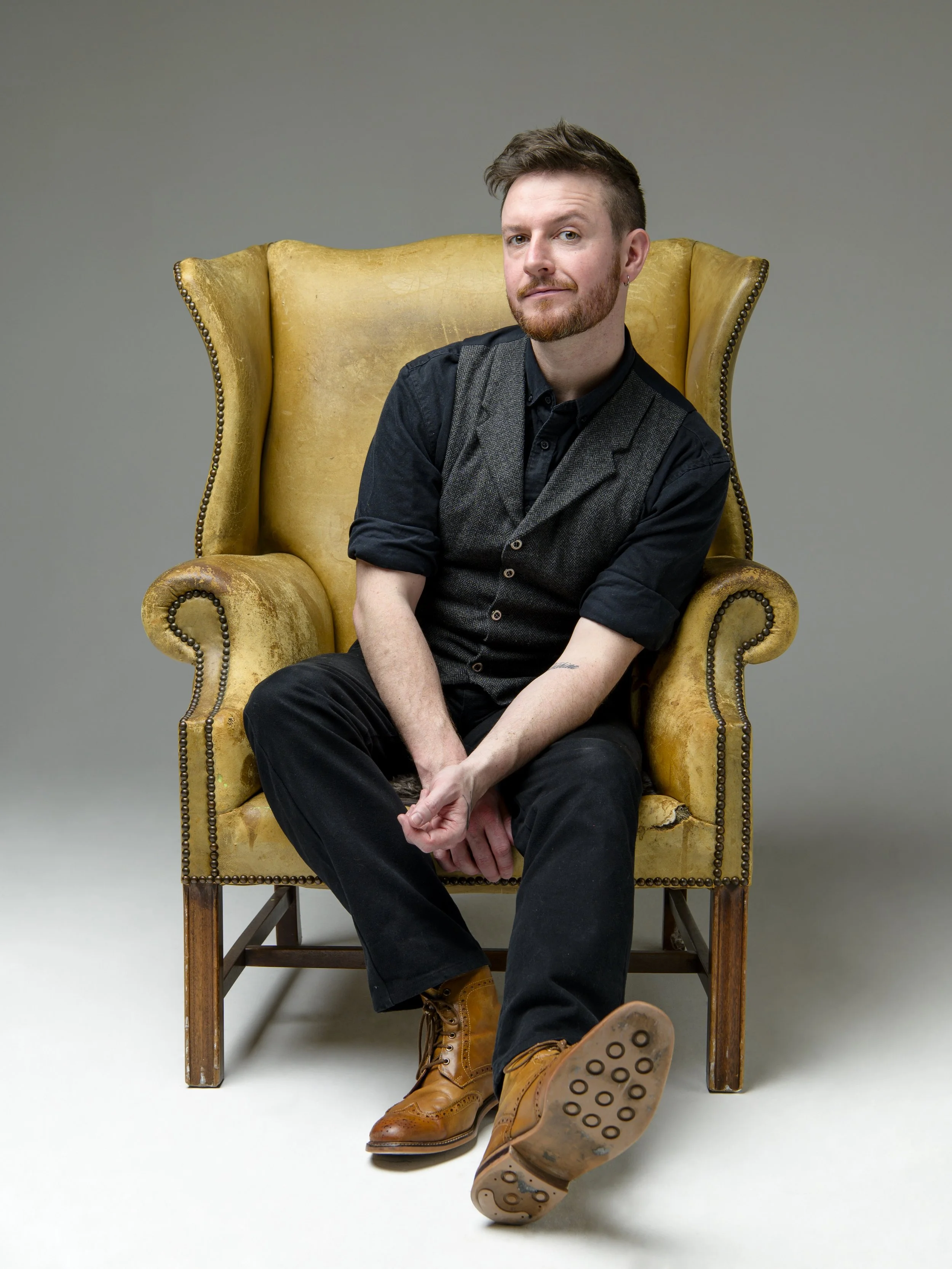 A young man with short brown hair, beard, and earring sitting on a yellow vintage armchair against a gray background. He is wearing a black shirt, gray vest, black pants, and tan boots, with a relaxed and confident expression.