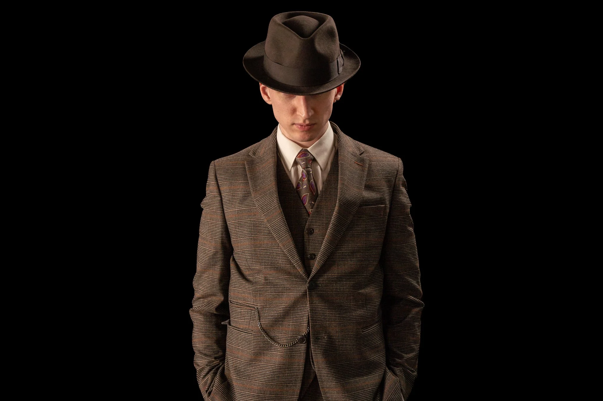 Young man in vintage-inspired brown plaid suit and tie, wearing a dark fedora, standing against a black background.