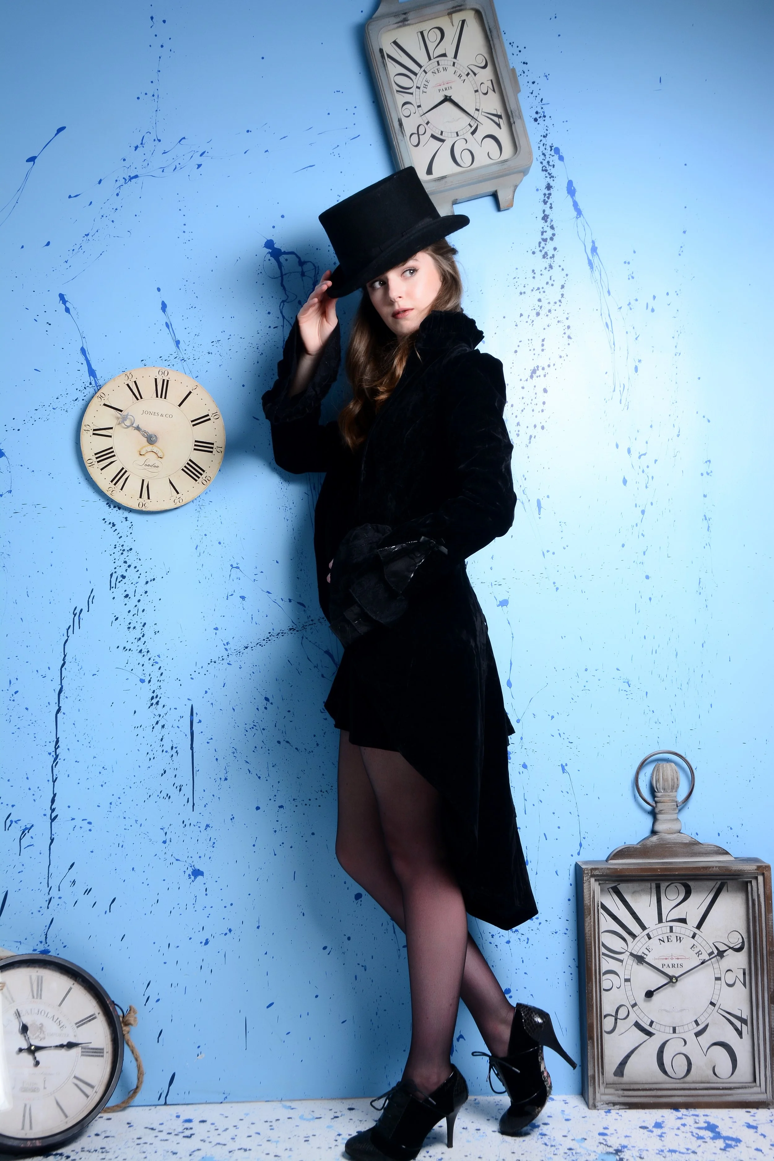 A young woman dressed in a black velvet coat, black high heels, and a top hat, stands against a blue wall with splattered paint. There are multiple vintage-style clocks on the wall and floor around her.