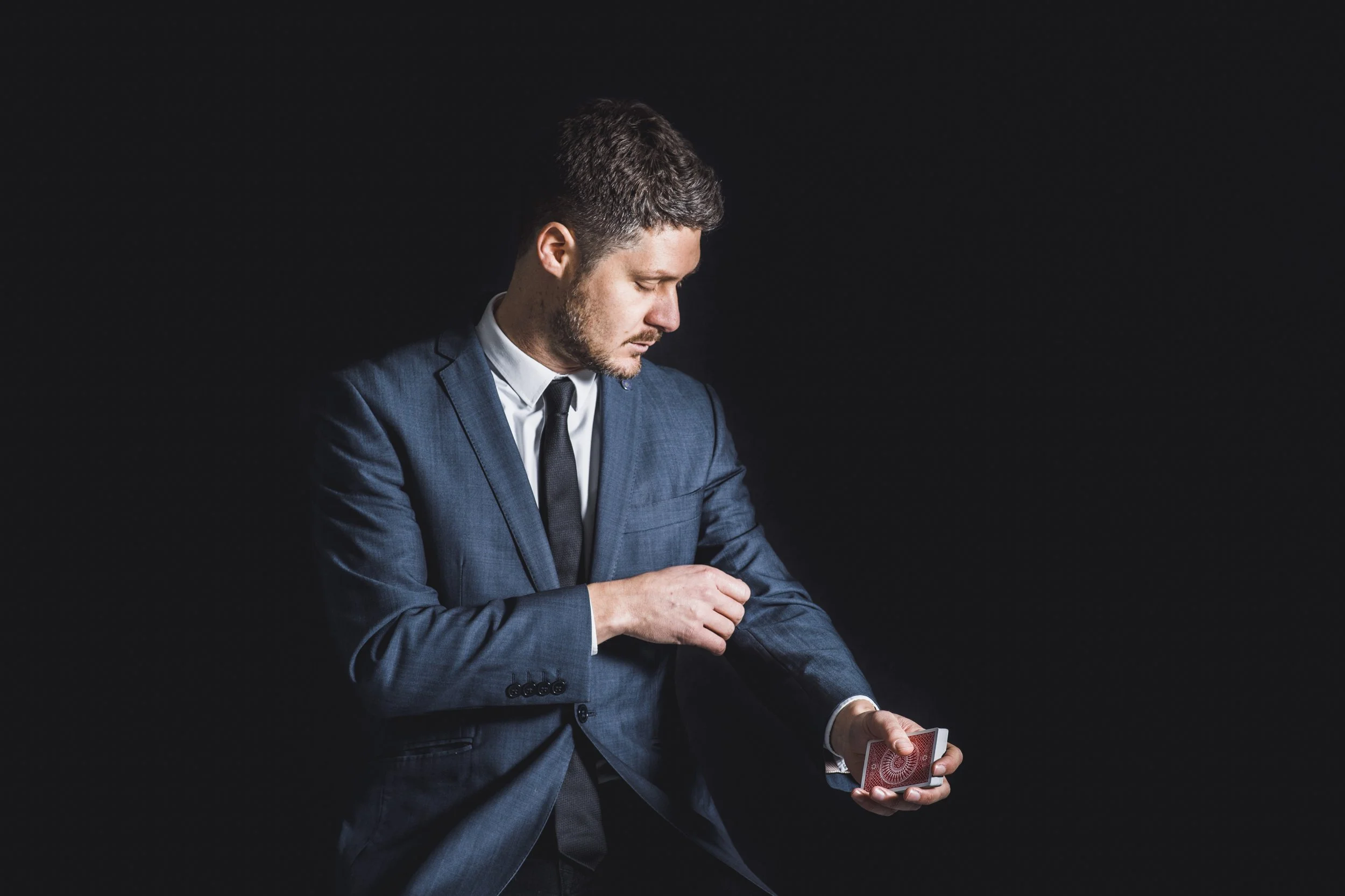 A man in a blue suit and black tie is looking at a card in his hand, with a black background.
