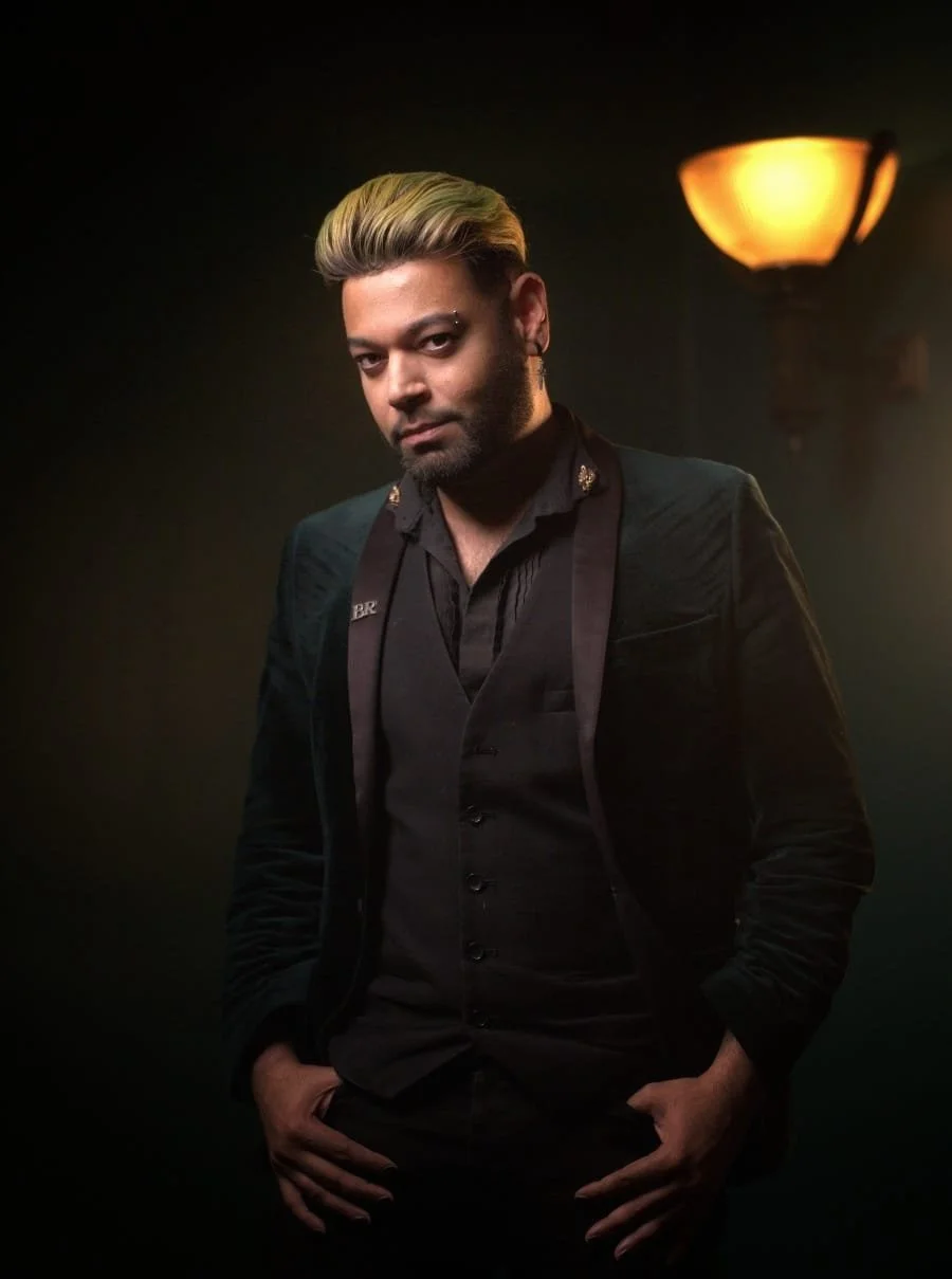 A man with blonde hair and a beard wearing a black suit and shirt, standing with hands in pockets in a dimly lit room with a warm wall lamp in the background.