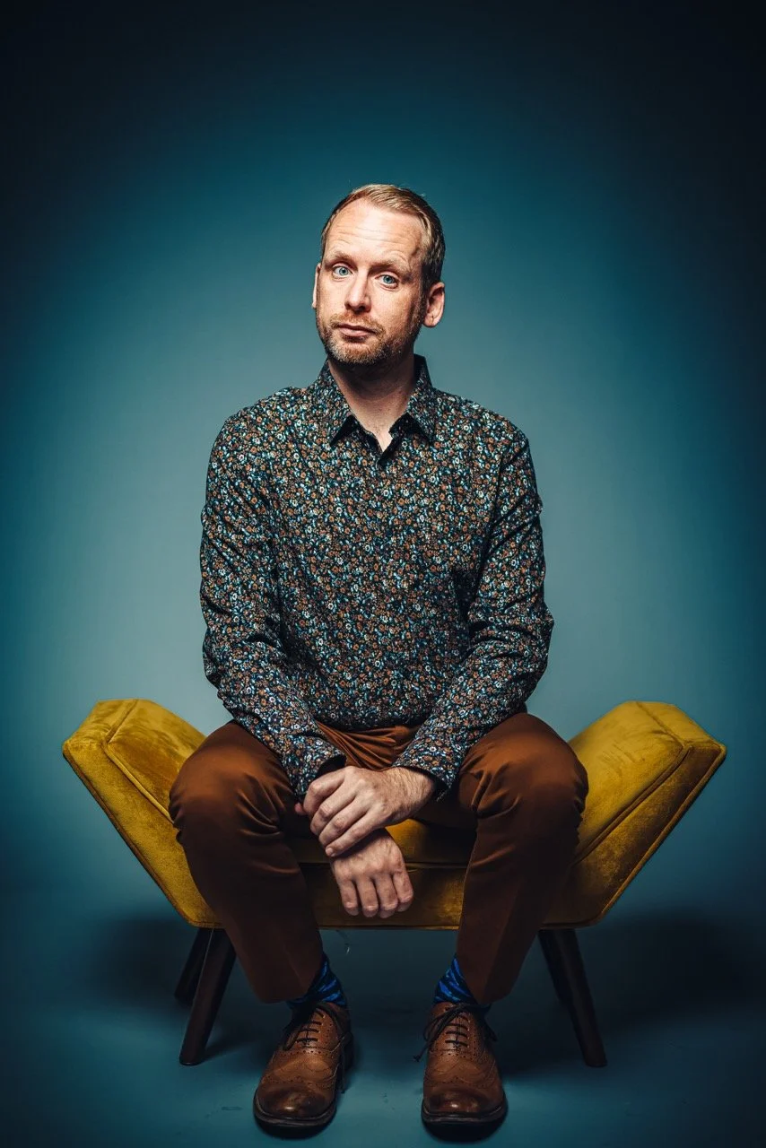 A man with light skin, reddish hair, and a beard sits on a yellow chair against a plain blue background. He wears a long-sleeve floral shirt, brown pants, blue patterned socks, and brown shoes, with a mildly inquisitive facial expression.
