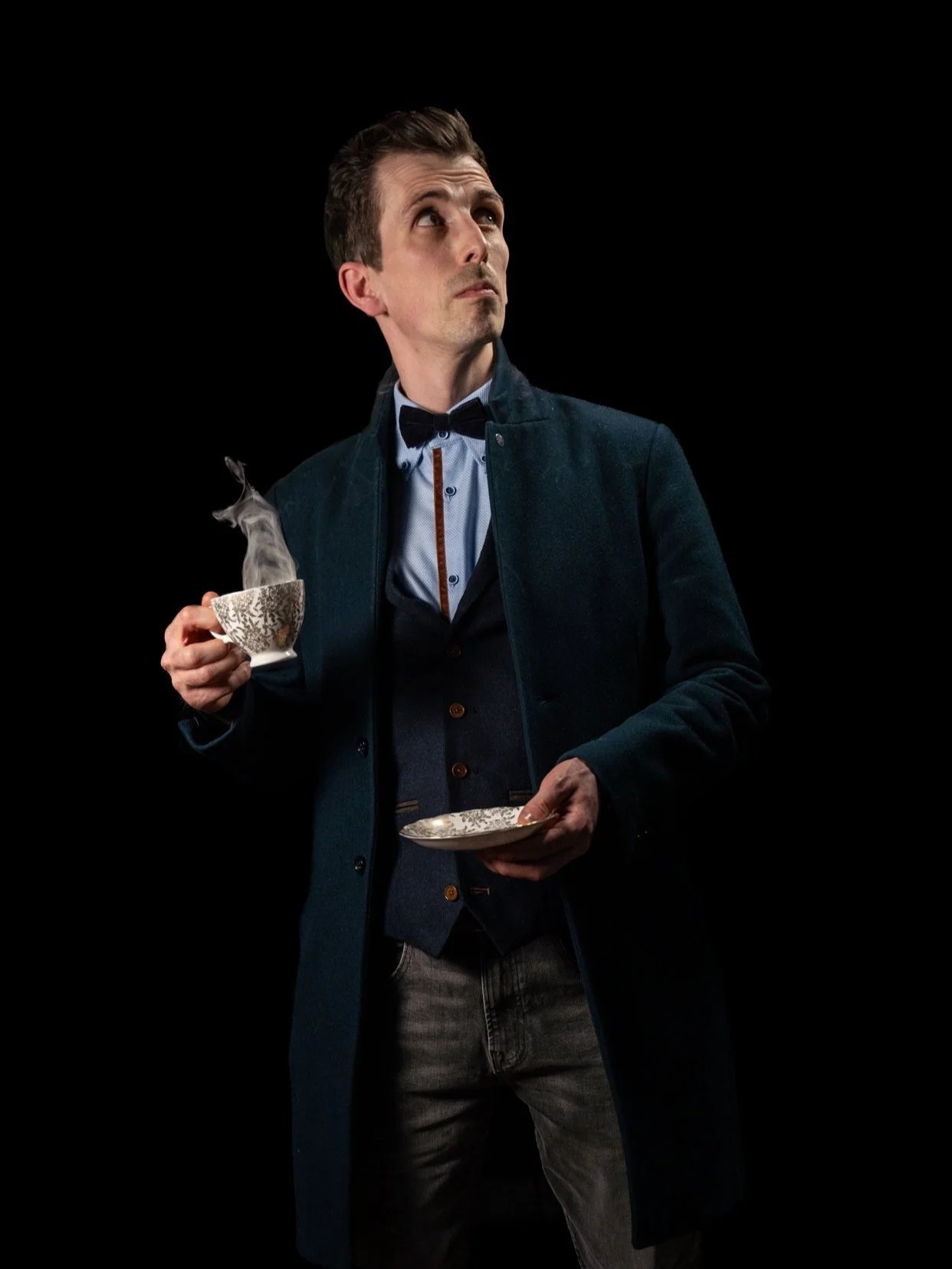 Chris Wilder wearing a dark coat, dress shirt, and bow tie holding a steaming cup and a matching plate against a black background.