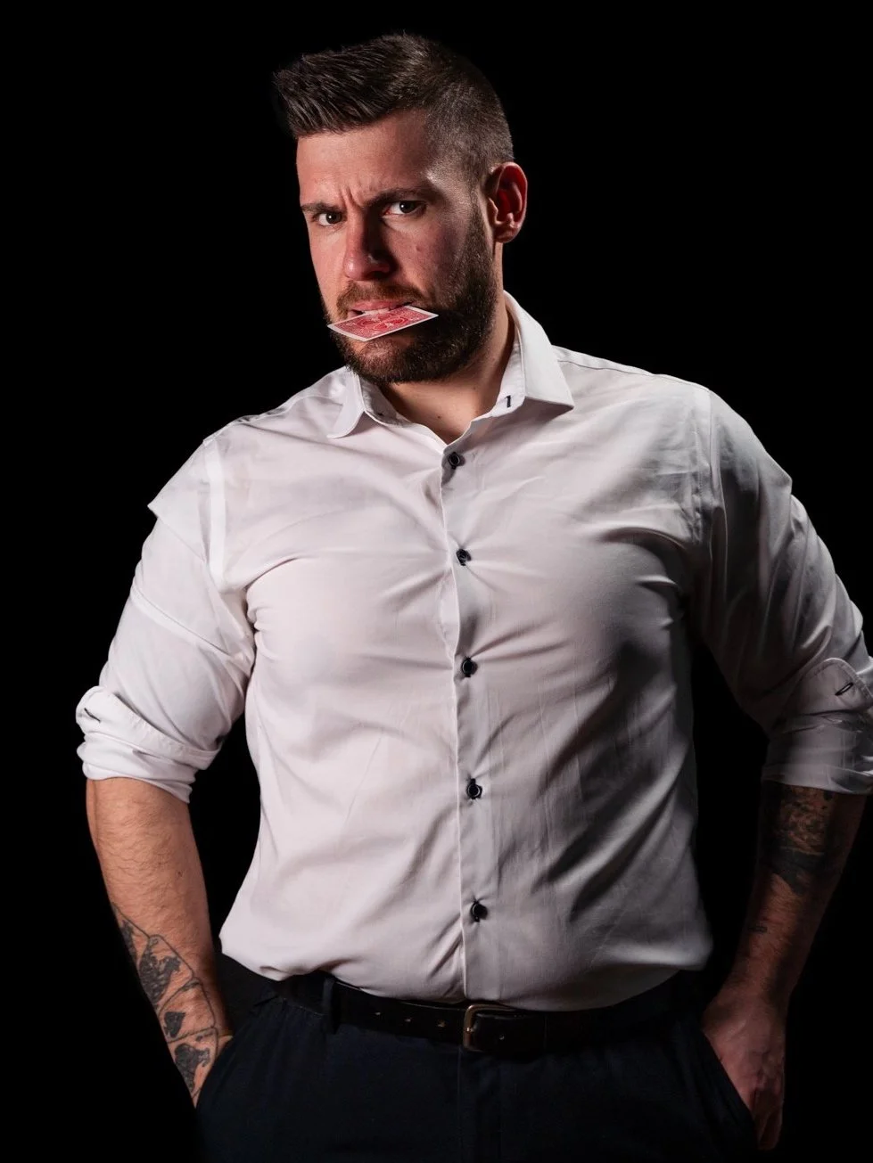 A man with a beard and tattoos on his arms, wearing a white dress shirt with rolled-up sleeves, holding a playing card in his mouth, against a black background.