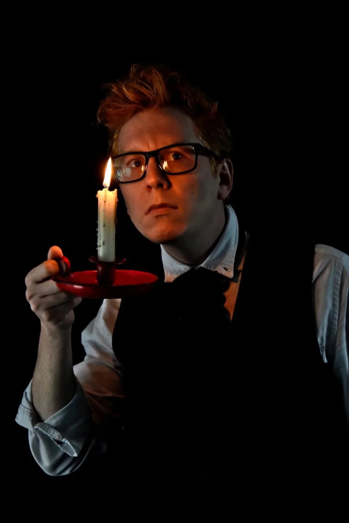 A person with red hair and glasses holding a lit candle on a red plate, looking intently into the distance against a black background.