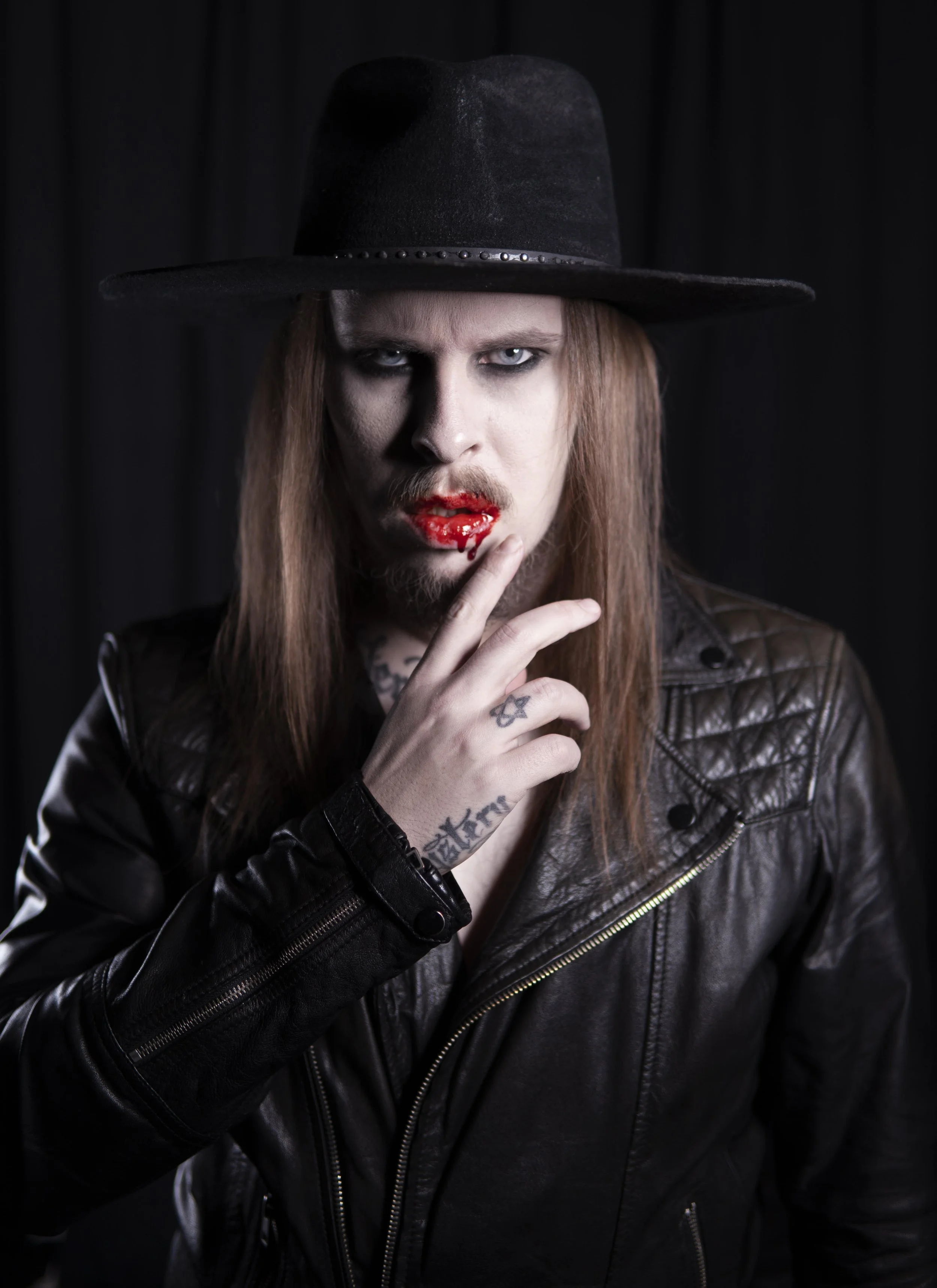 A person with long red hair wearing a black wide-brimmed hat and black leather jacket, with makeup and blood on their lips, holding their finger near their lips, with tattoos visible on their hand and neck.