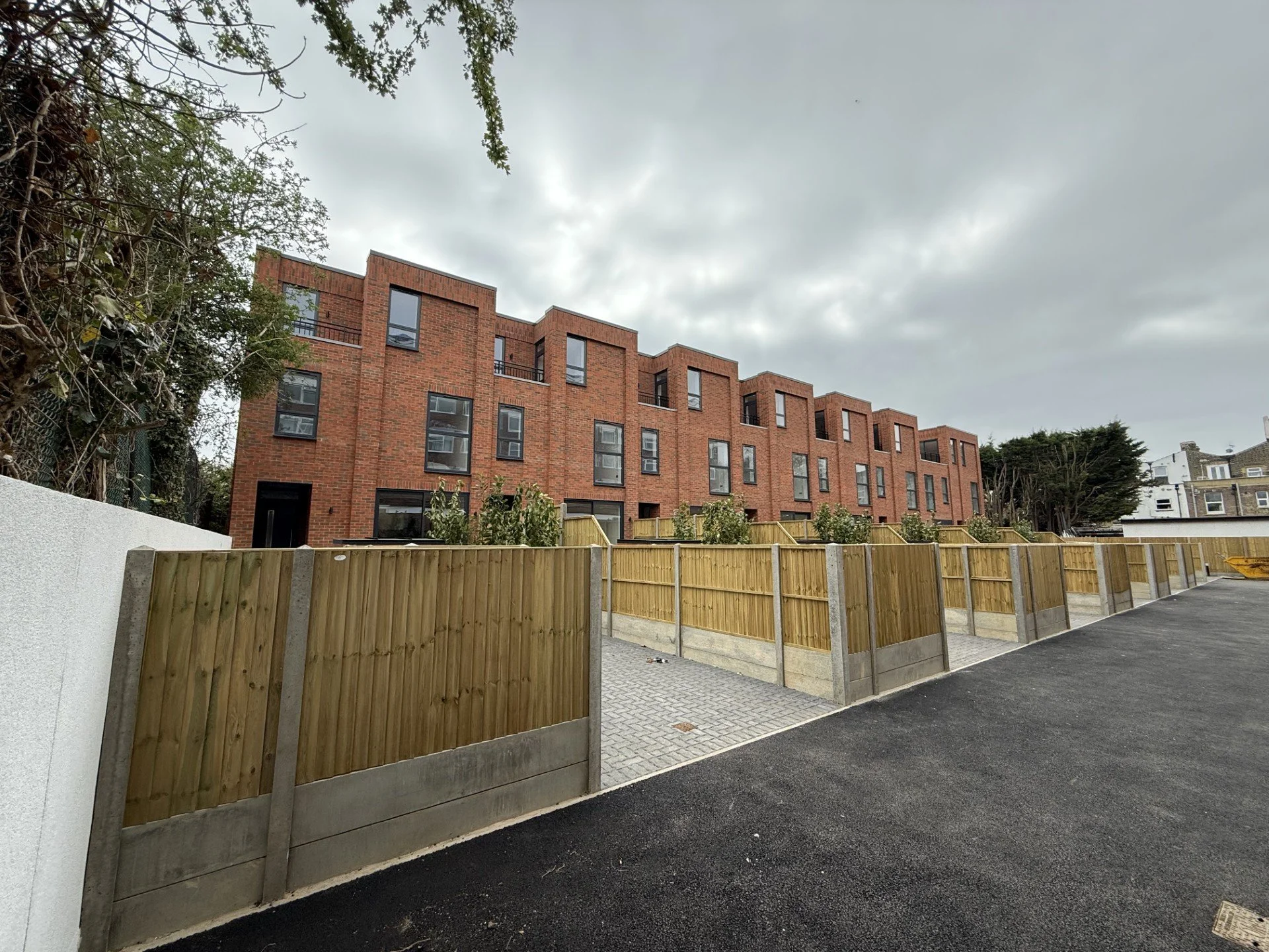 Newly constructed red brick apartment building with fenced yard and small trees, under a cloudy sky.