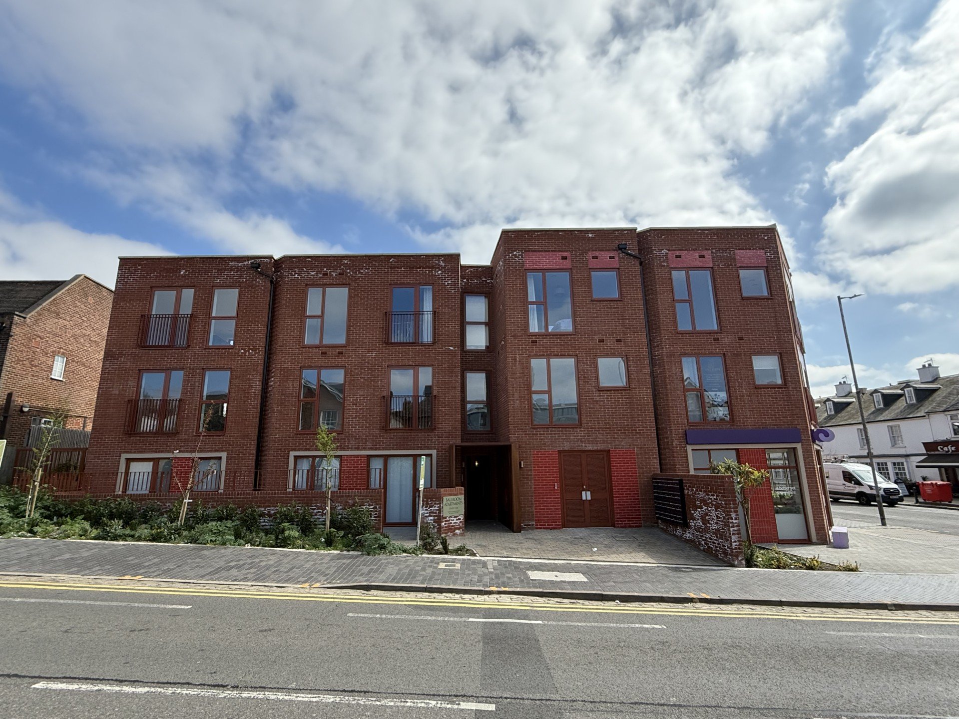 Red brick apartment building with four stories, large windows, and small balconies on some units, set against a partly cloudy sky.