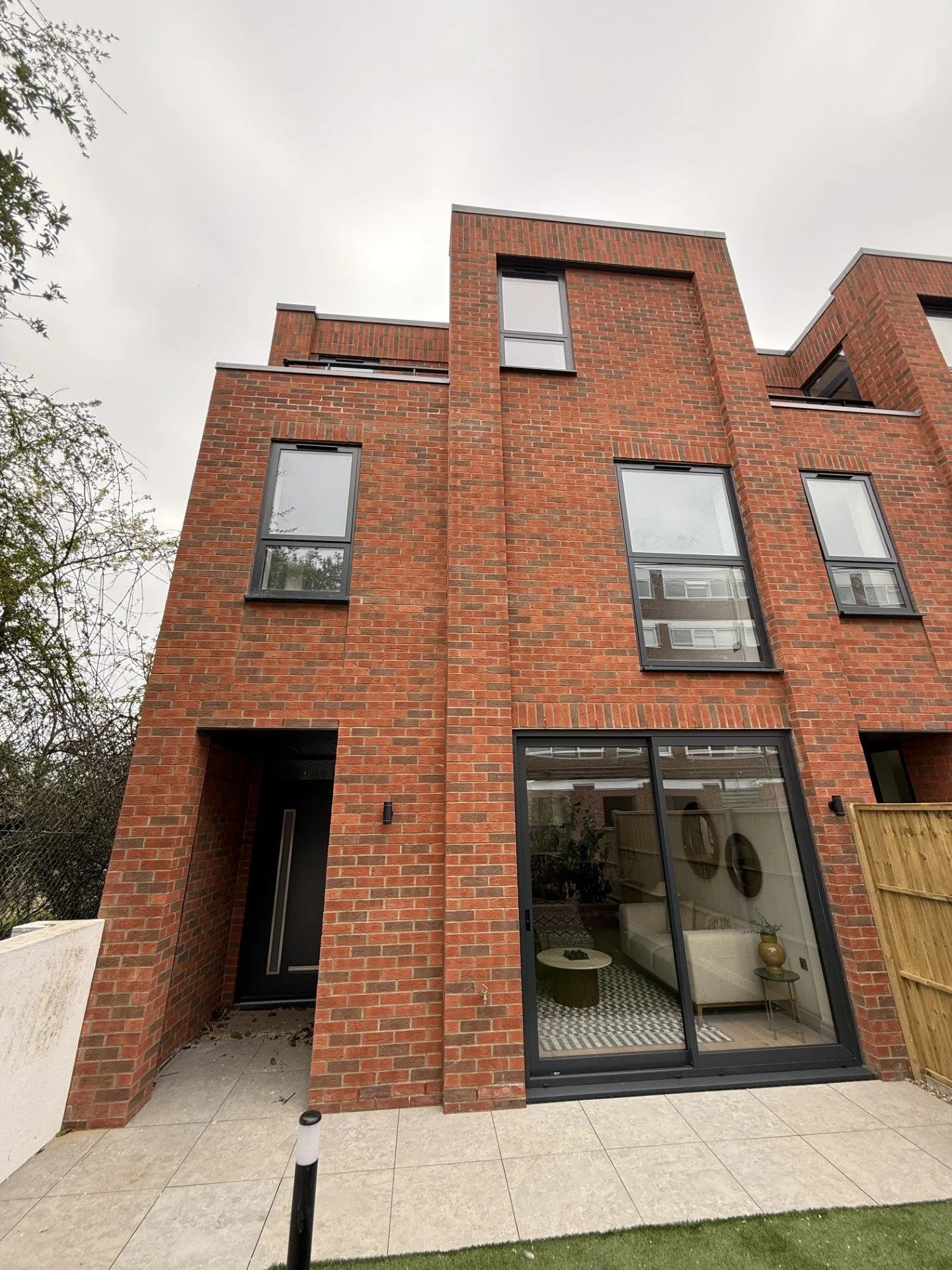 The image shows the exterior of a modern, multi-story brick building with several large windows, a sliding glass door revealing an interior living space, and a small patio area with tiled flooring and a grassy area.