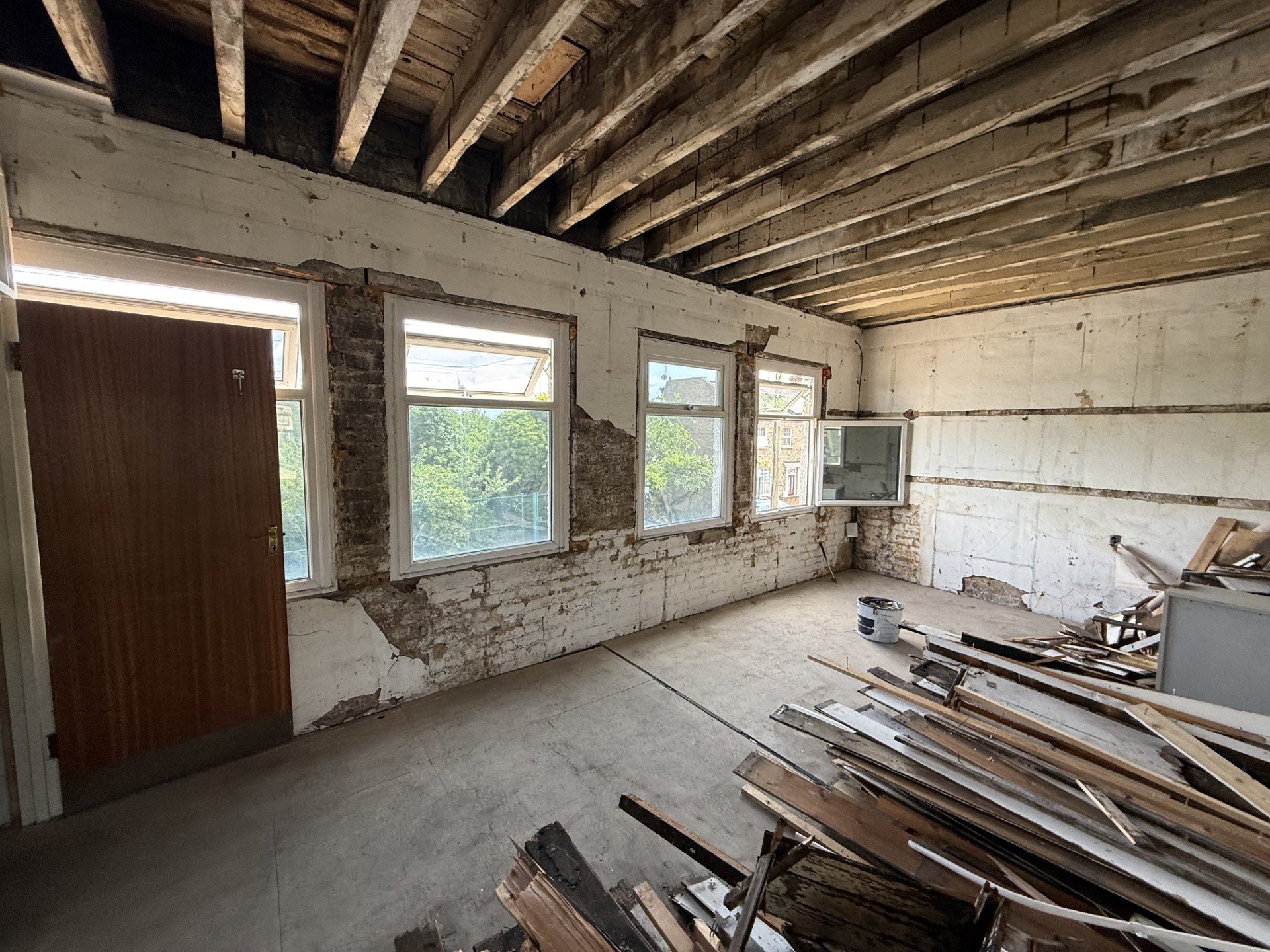 Interior of a room under renovation with exposed brick walls, large windows, and a wooden ceiling; construction materials and debris on the floor.