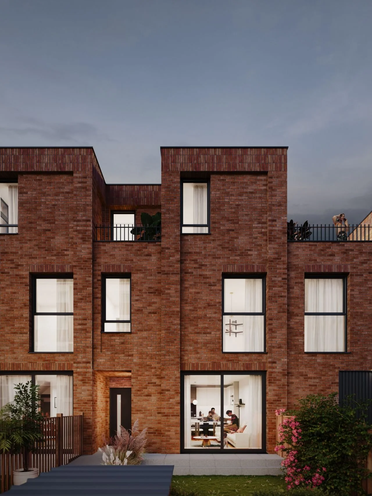 A multi-story red brick residential building seen from the backyard, with large windows showing interior living spaces, plants, and outdoor furniture.