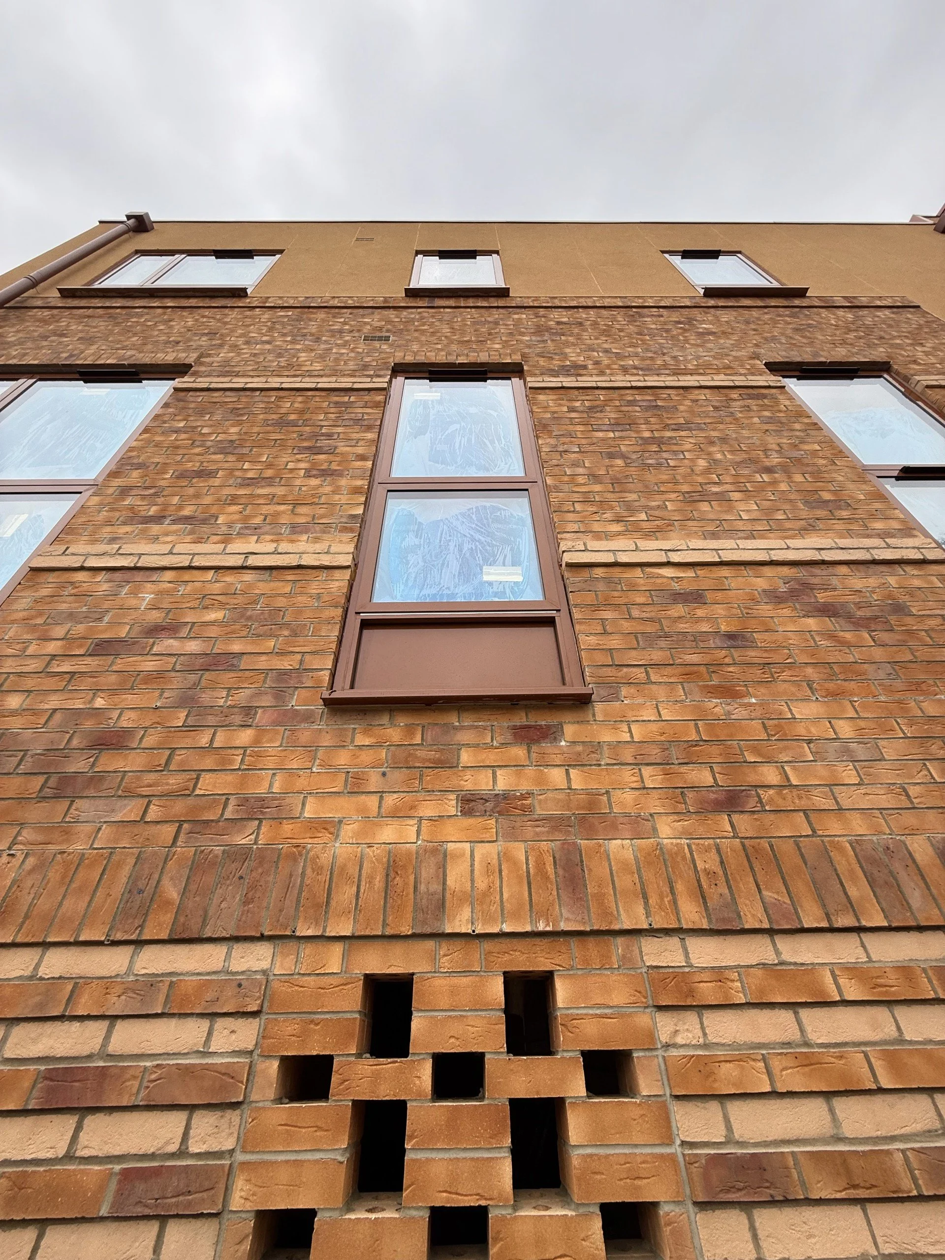 Looking up at a brick building with multiple windows and a vent at the bottom, overcast sky in background.