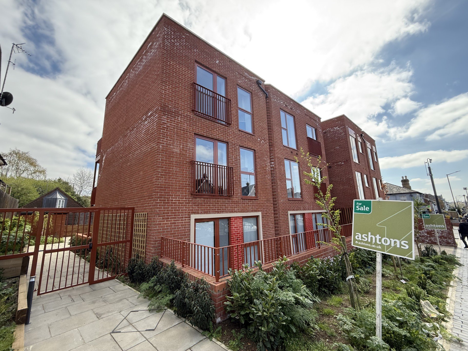 Brick apartment building with multiple windows and balconies, for sale sign in front, sidewalk with plants, cloudy sky in the background.