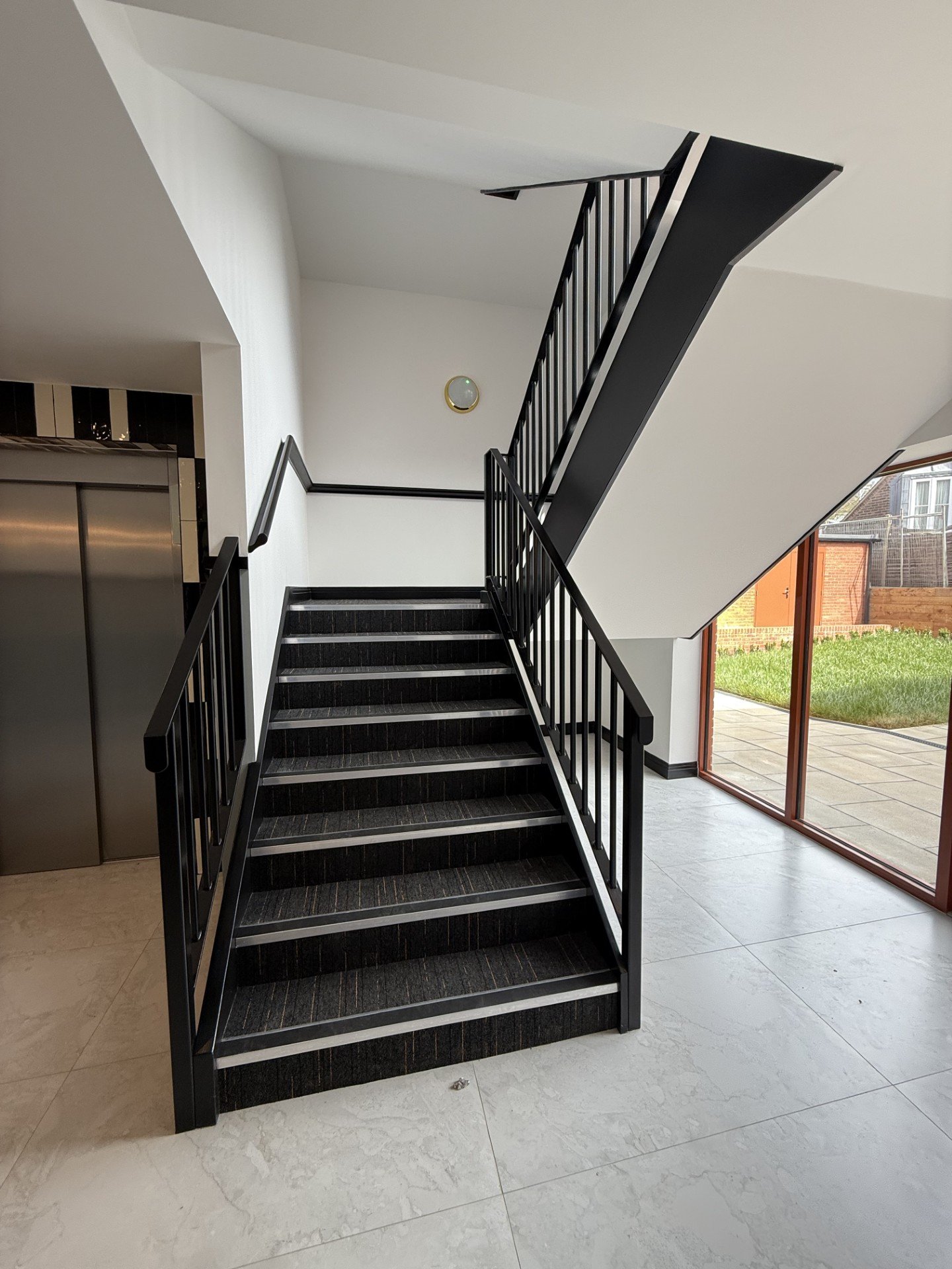 Indoor staircase with black steps and black railings, leading to an upper level. Large glass sliding door showing a grassy outdoor area.