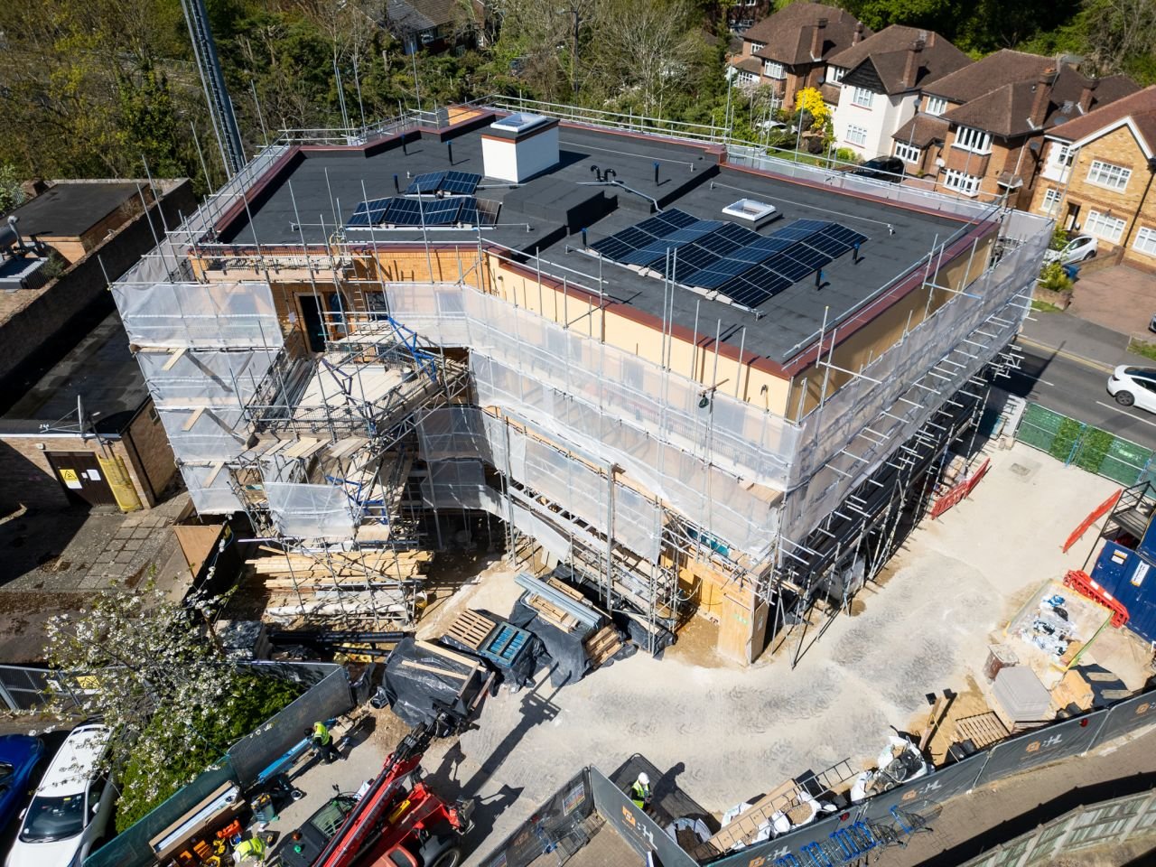 Aerial view of a building under construction with scaffolding, solar panels on the roof, and construction workers at the site.