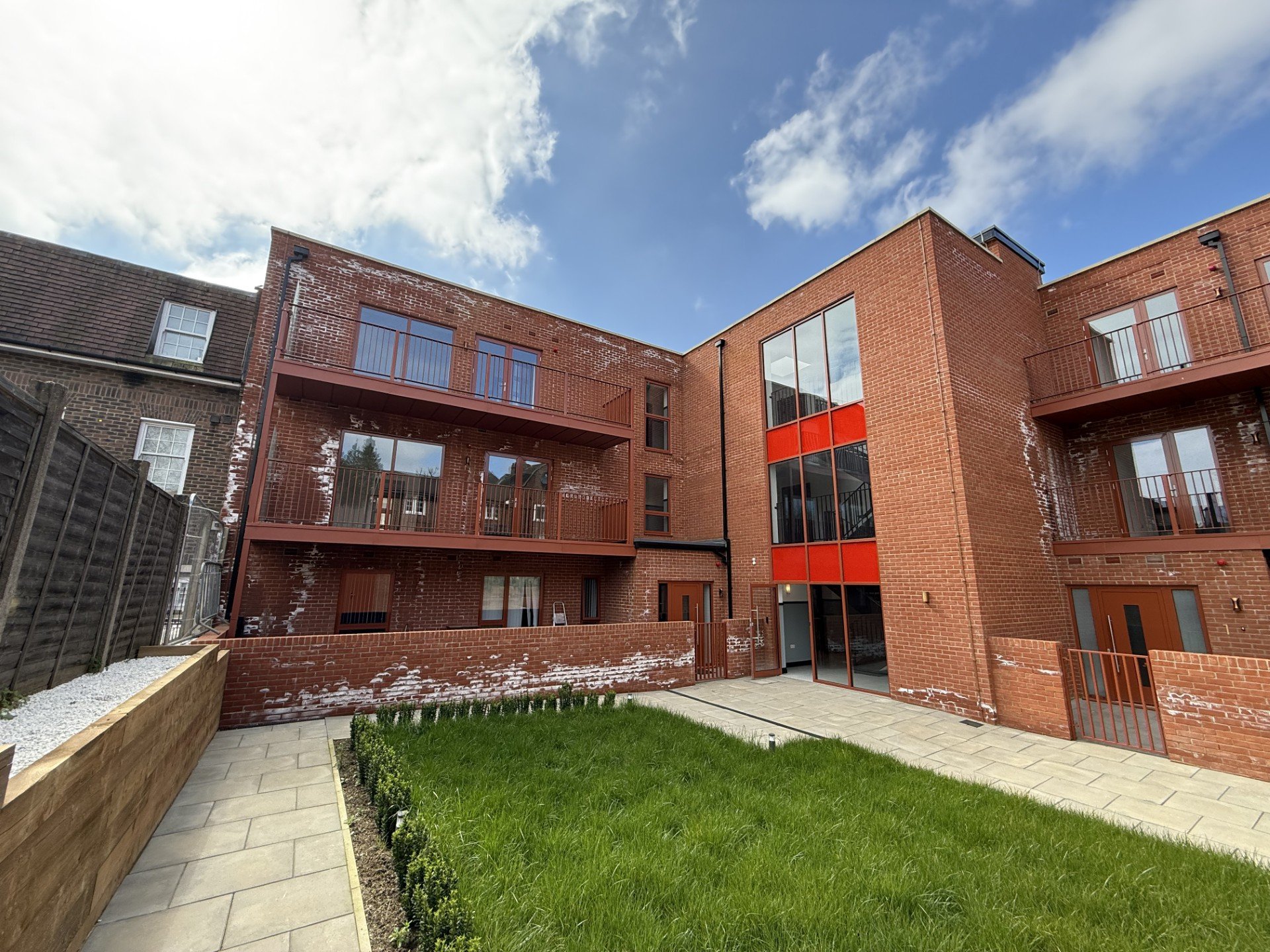 Modern red brick apartment building with balconies, glass doors, and a small courtyard with green grass, paved walkway, and a wooden planter. Blue sky with clouds overhead.