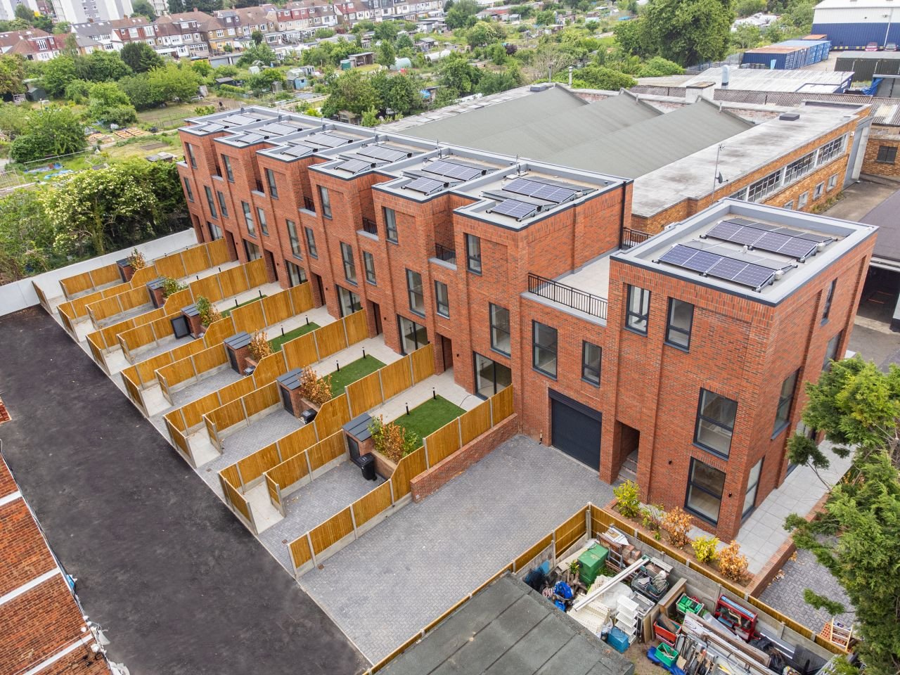 Aerial view of modern brick townhouses with individual fenced backyards, solar panels on rooftops, and a large paved driveway in a residential neighborhood.