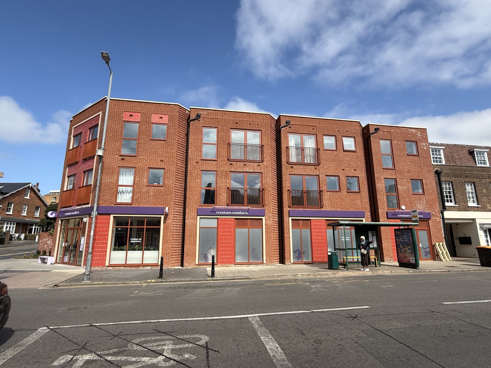 A multi-story brick building with a storefront labeled `creature comforts` on the ground floor, a bus stop with a person waiting, and a street in the foreground with parking markings. The sky is partly cloudy.