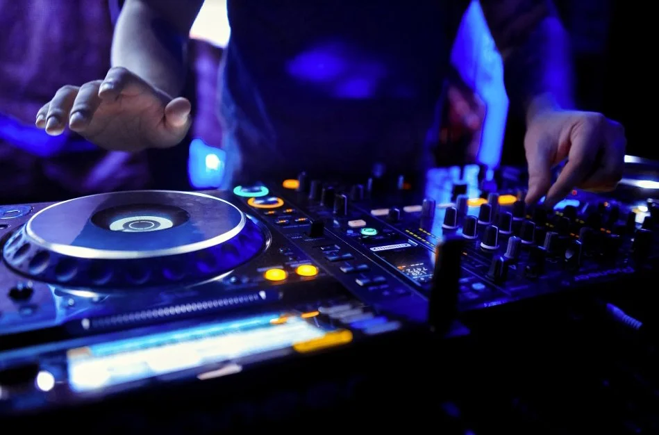 Close-up of a DJ manipulating a professional DJ mixer with illuminated controls in a dark, nightclub setting.