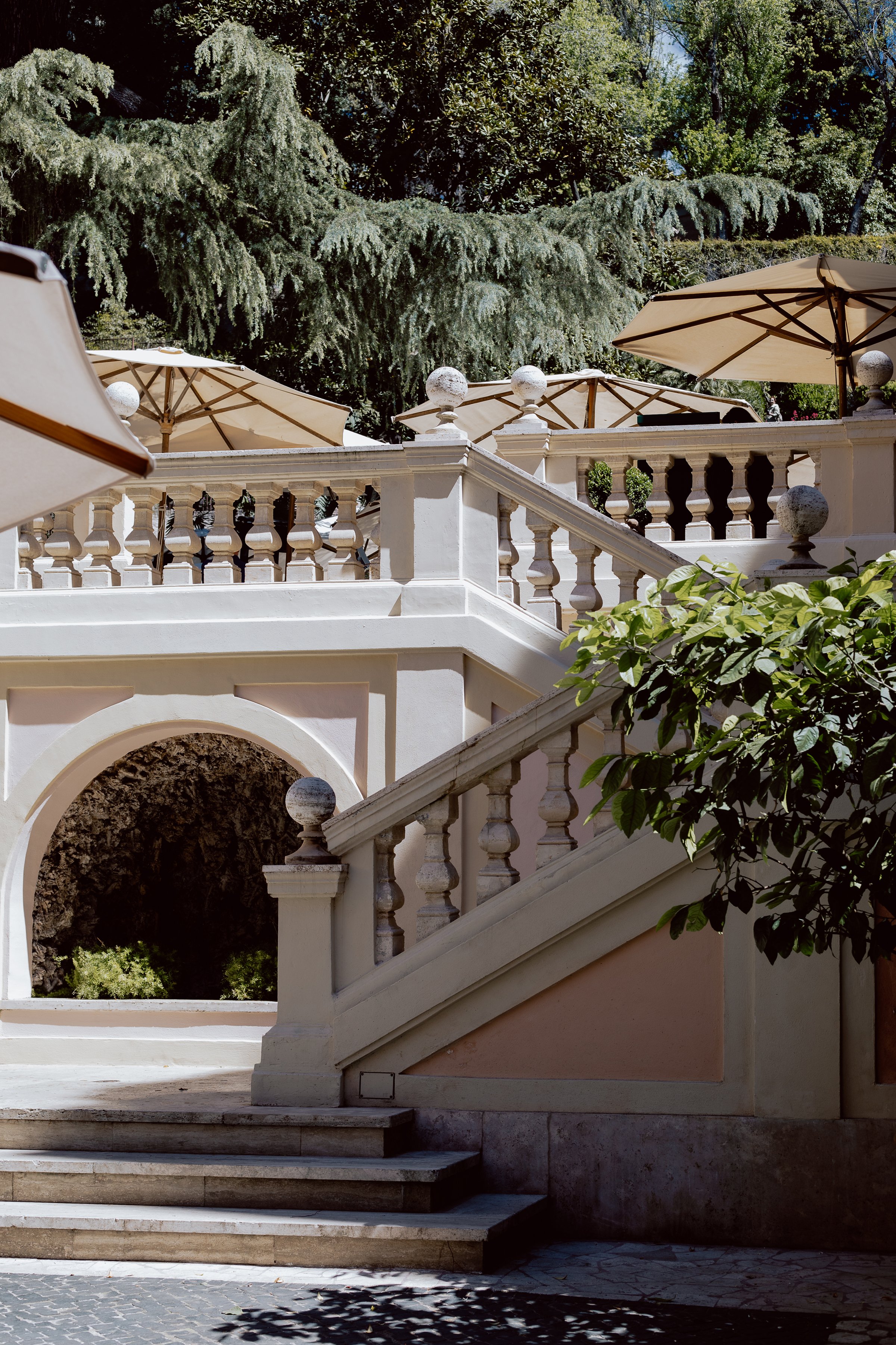 Outdoor staircase with white balustrades and steps, surrounded by green trees and plants, with umbrellas shading a terrace area.
