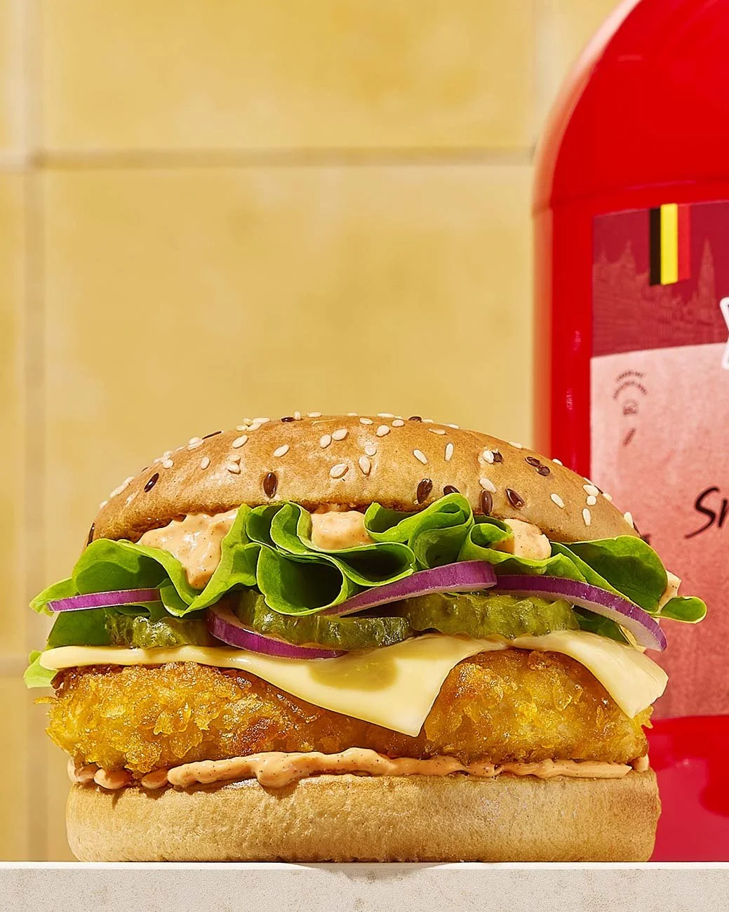 Food and tabeltop styling Gent. A crispy chicken sandwich with lettuce, pickles, red onion, cheese, and sandwich sauce in a sesame seed bun