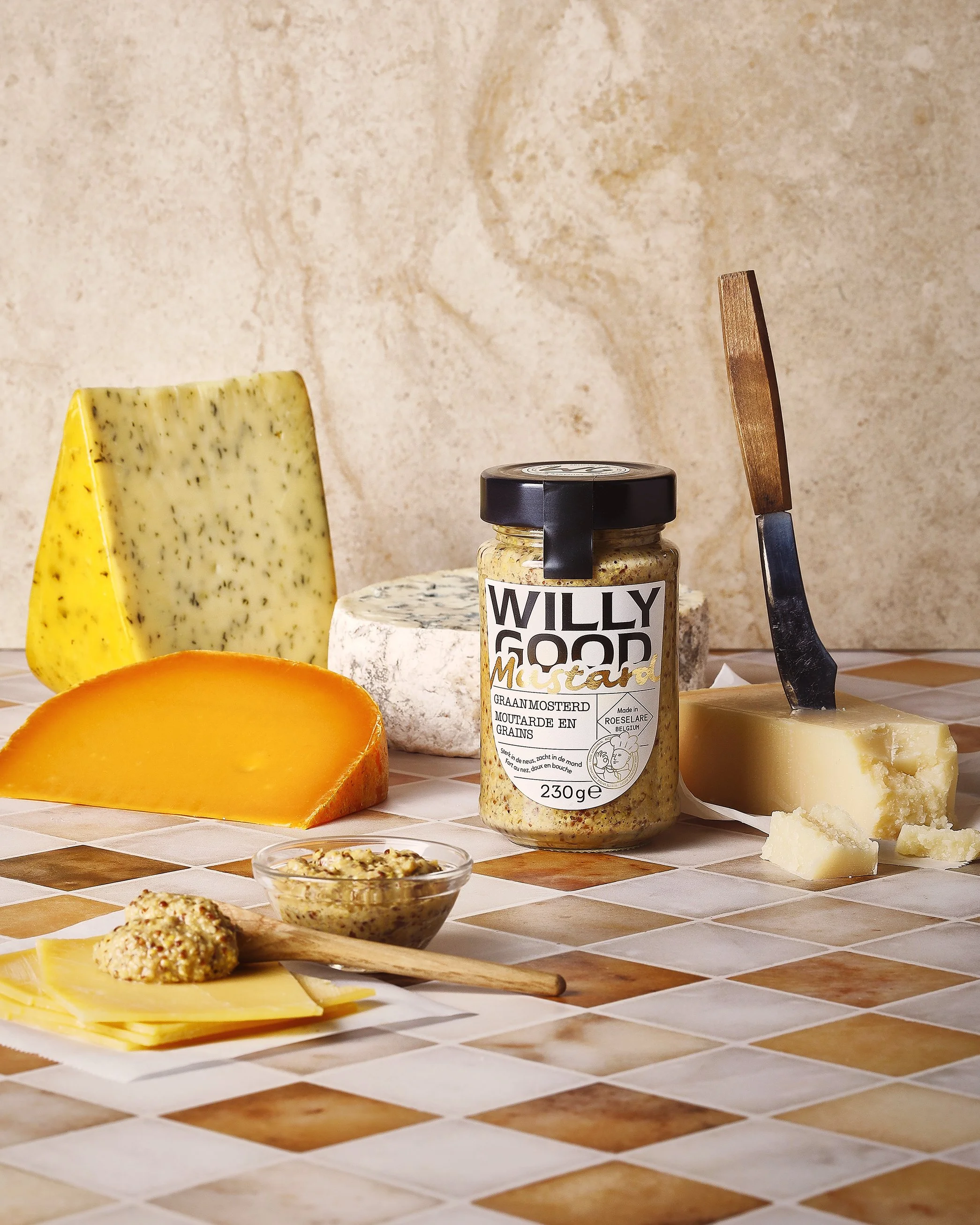 Food styling, tabletop styling and art direction Willy Good, Gent, Various cheeses, a jar of mustard, and a knife arranged on a tiled surface, with a light beige textured background.