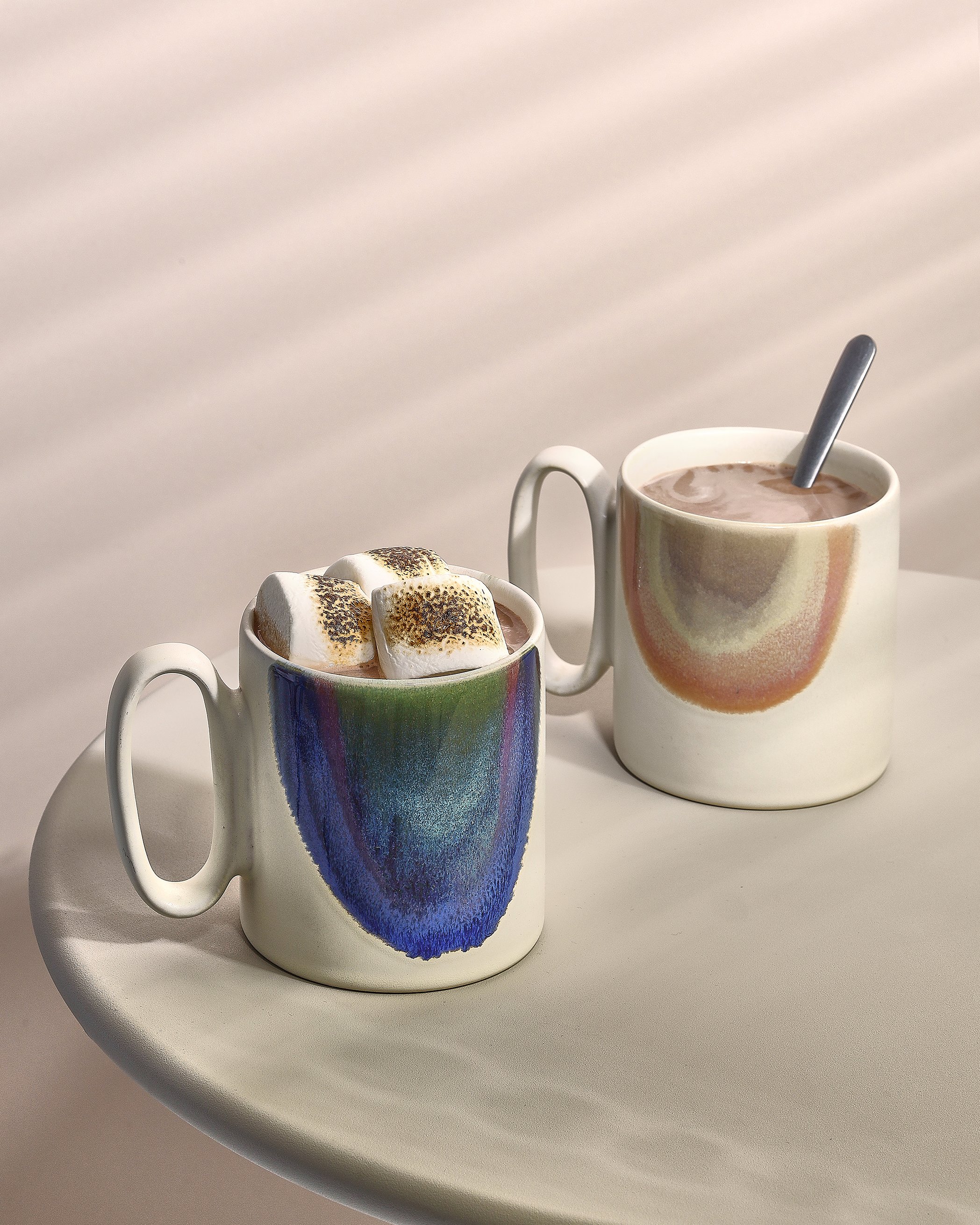 Sarah Ghijselinck food stylist, Two ceramic Salt&pepper mugs with colorful abstract paintings on them, one filled with hot chocolate topped with marshmallows and the other with hot chocolate.