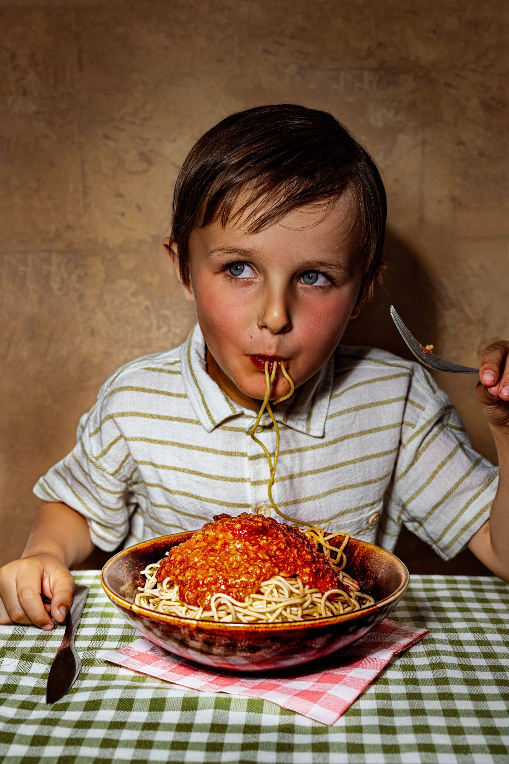 Sarah Ghijselinck, food and drink stylist, art direction, recipe development, Gent, fine dining, restaurant photography for levi's burgers. A young boy is eating spaghetti with tomato sauce at a table with a green checkered tablecloth.