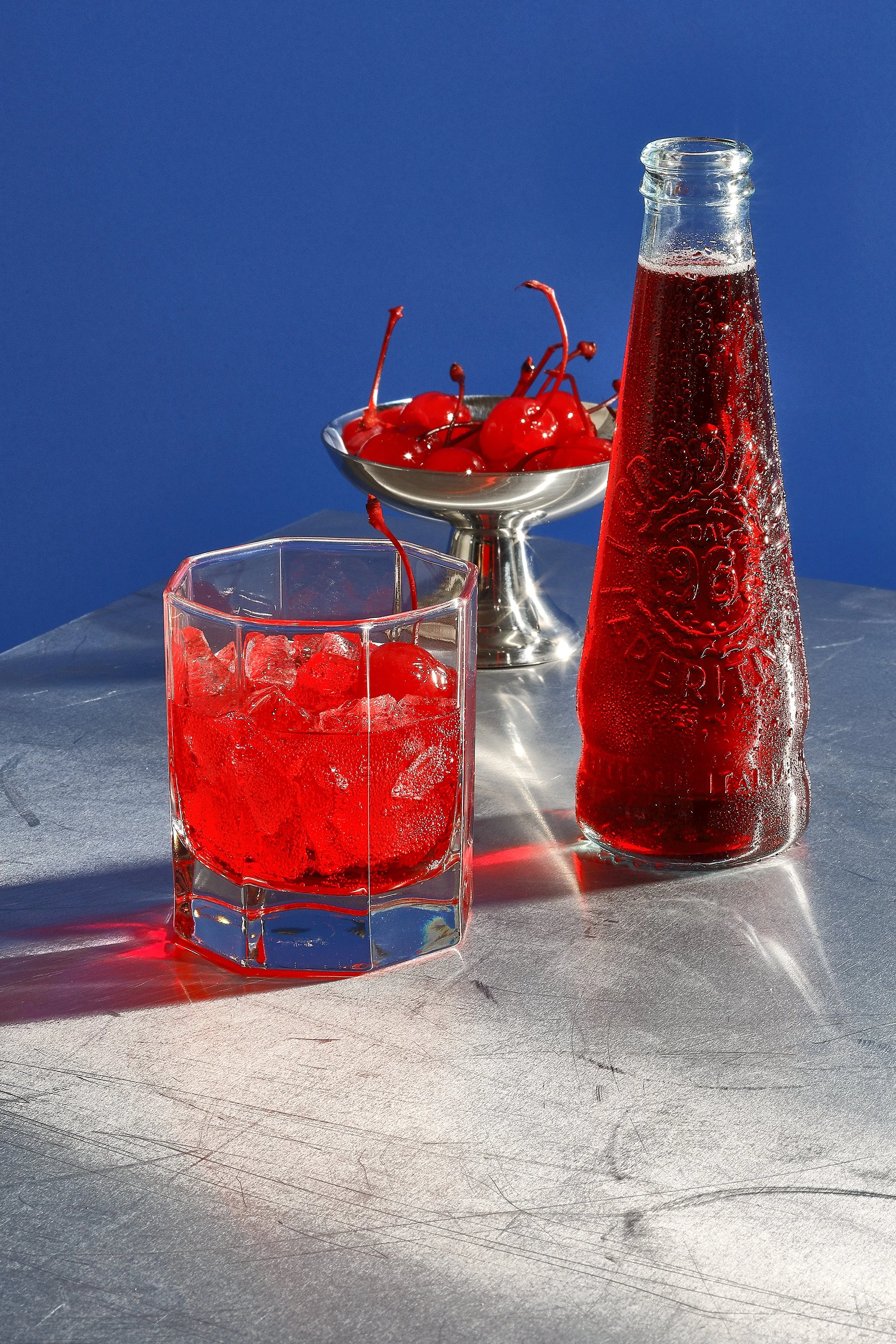 Drink styling Gent. A glass of cherry-flavored soda with ice, a metal dish of cherries, and a glass bottle of cherry soda on a metallic surface against a blue background.