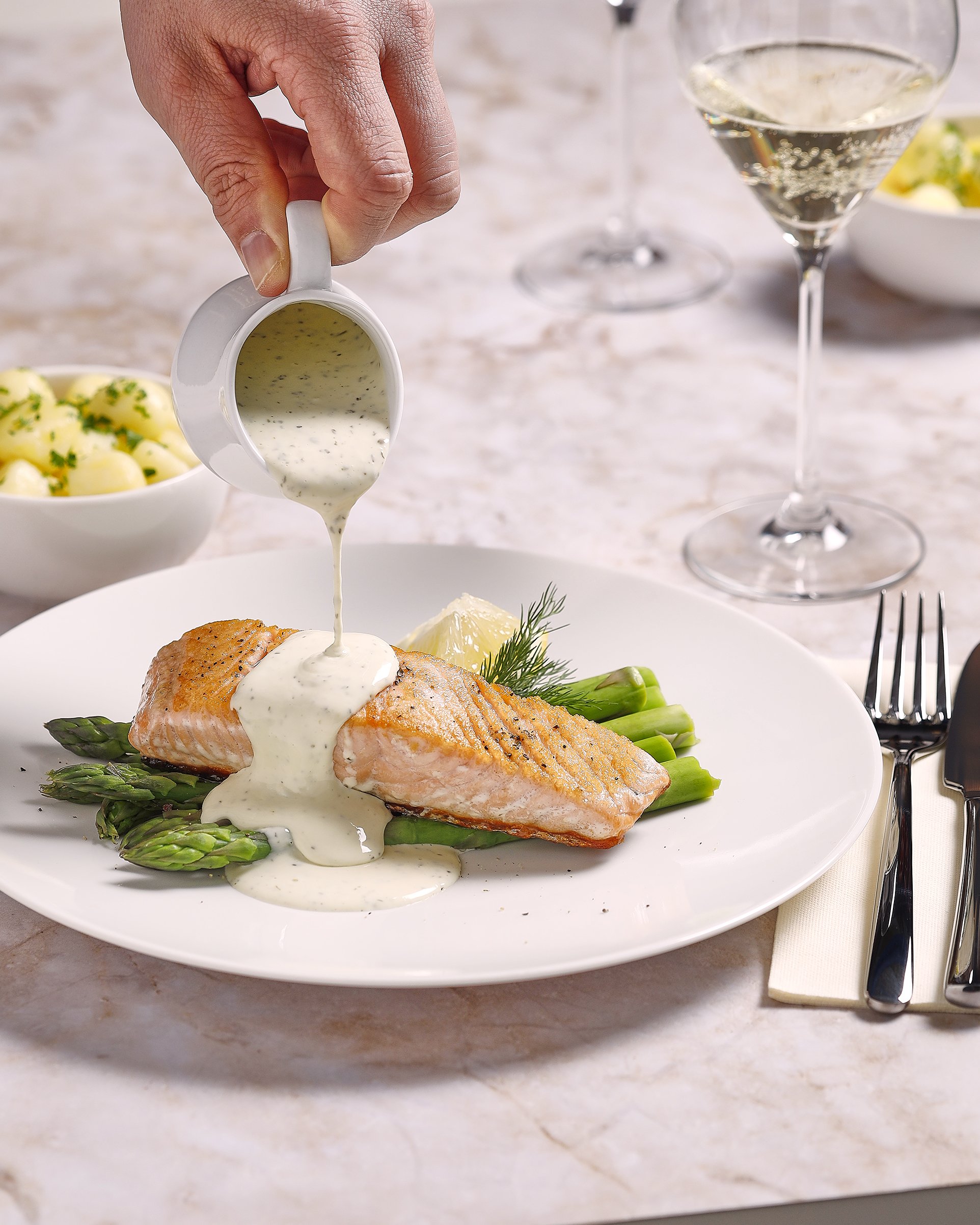 Sarah Ghijselinck food stylist, tabletop styling. restaurant fine dining, A plate with a piece of cooked salmon, asparagus, a lemon wedge, and fresh herbs, with sauce being poured over the salmon. 