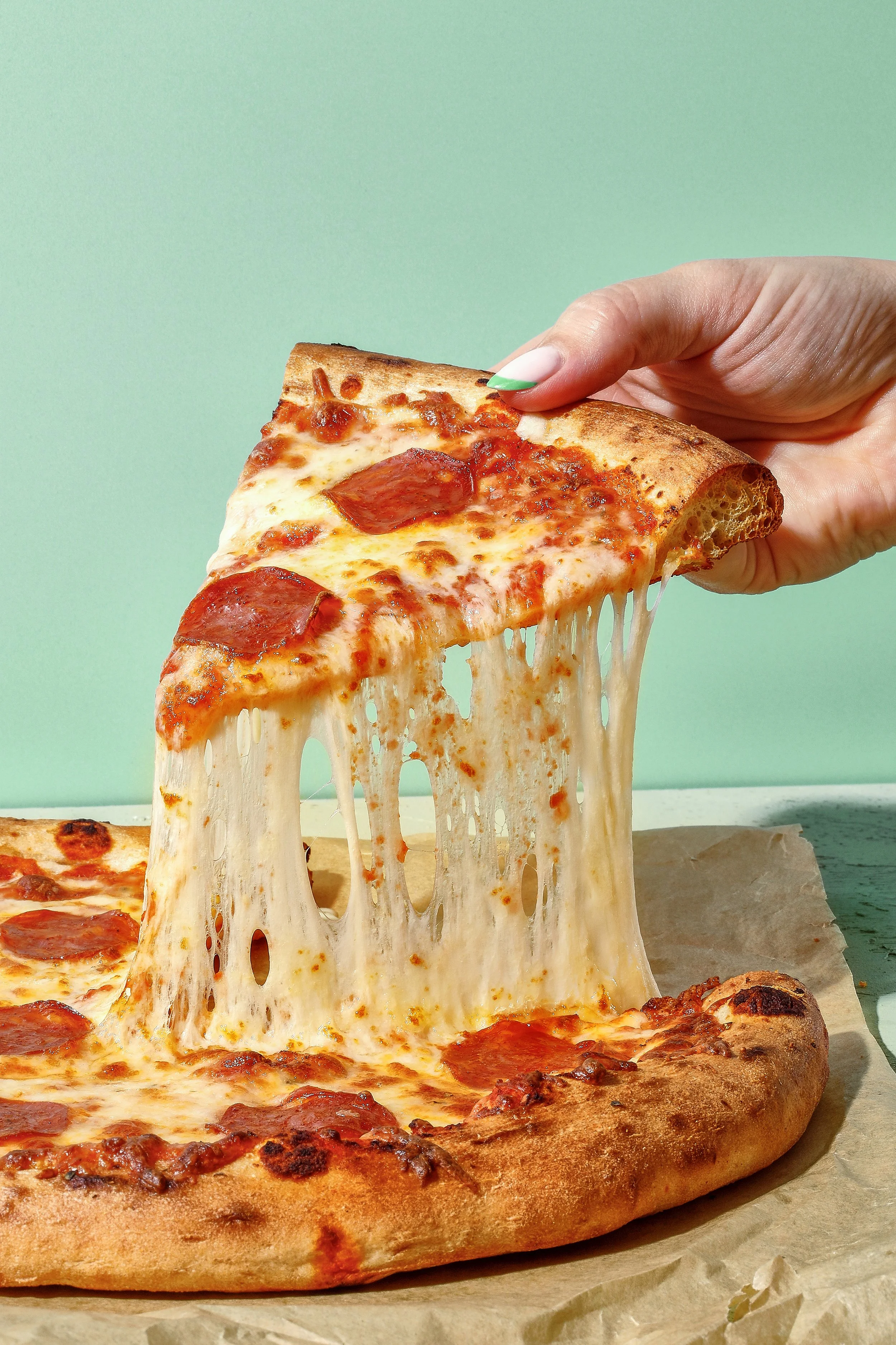 Sarah Ghijselinck, food and drink stylist, Gent, art direction, A hand lifting a slice of pepperoni pizza with melted cheese stretching from the pizza. 
