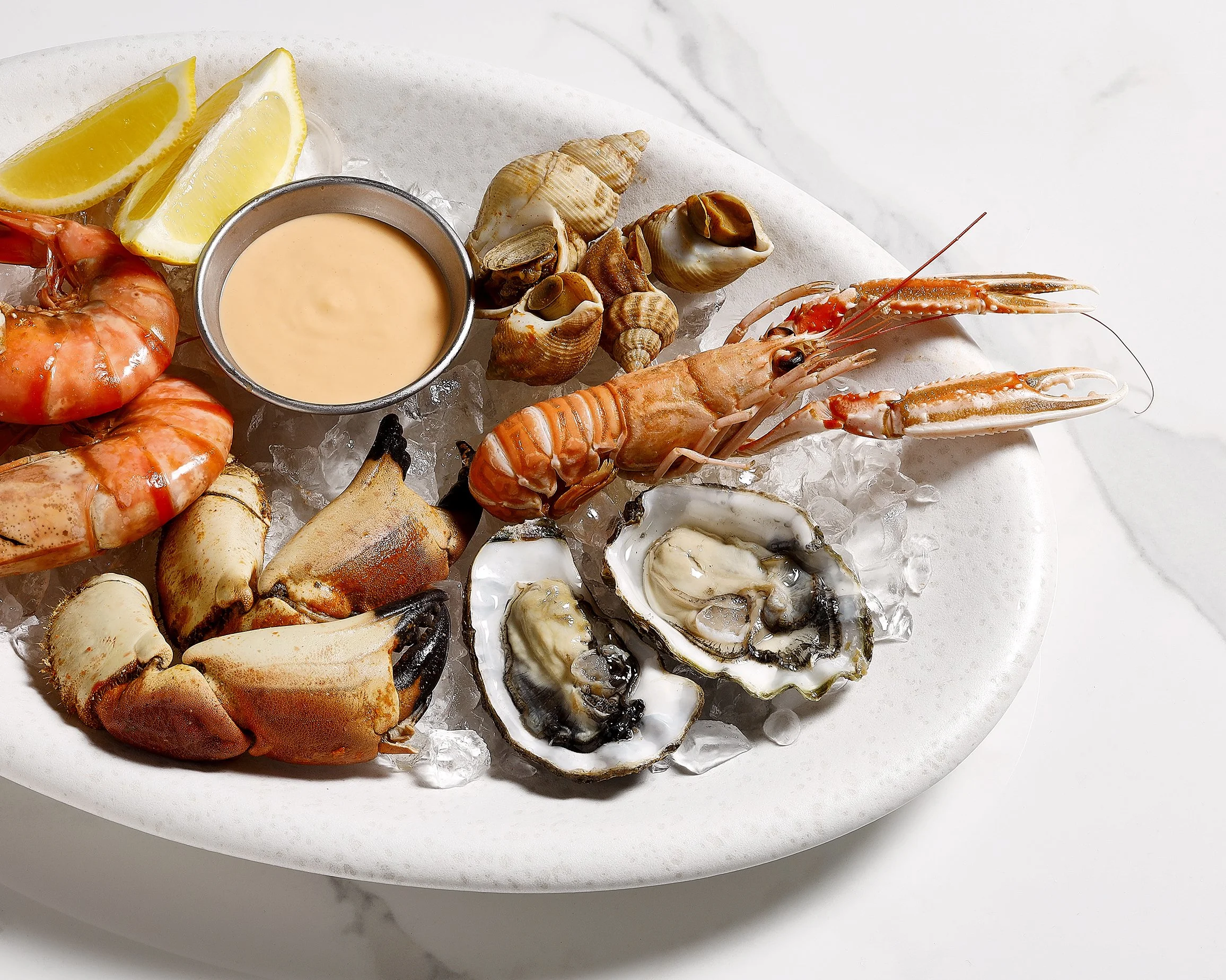 Sarah Ghijselinck food stylist, Seafood platter with lobster, shrimp, oysters, clams, and a dipping sauce, garnished with lemon wedges, on a white plate.