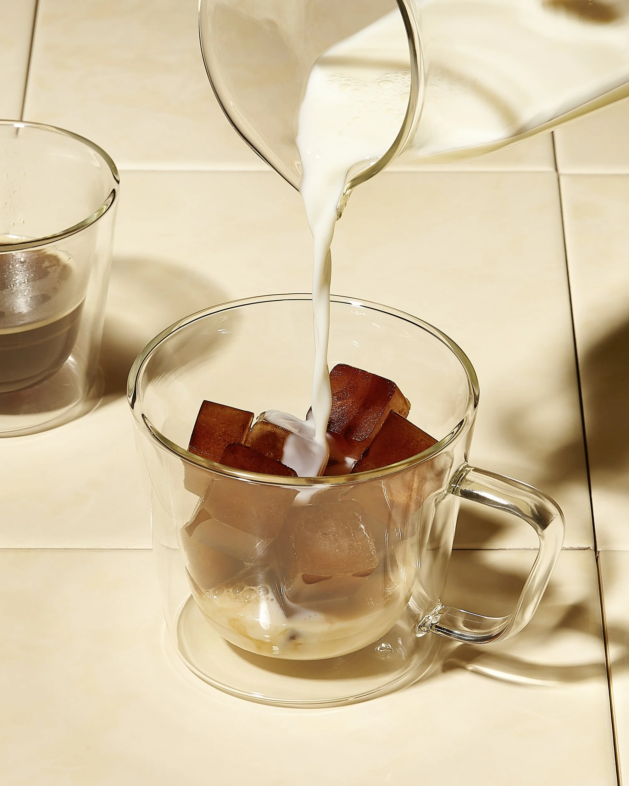 Sarah Ghijselinck, food and drink stylist, art direction, recipe development, Gent, Cream is being poured into a clear glass mug from salt&pepper with brown sugar cubes, on a light-colored tiled surface.