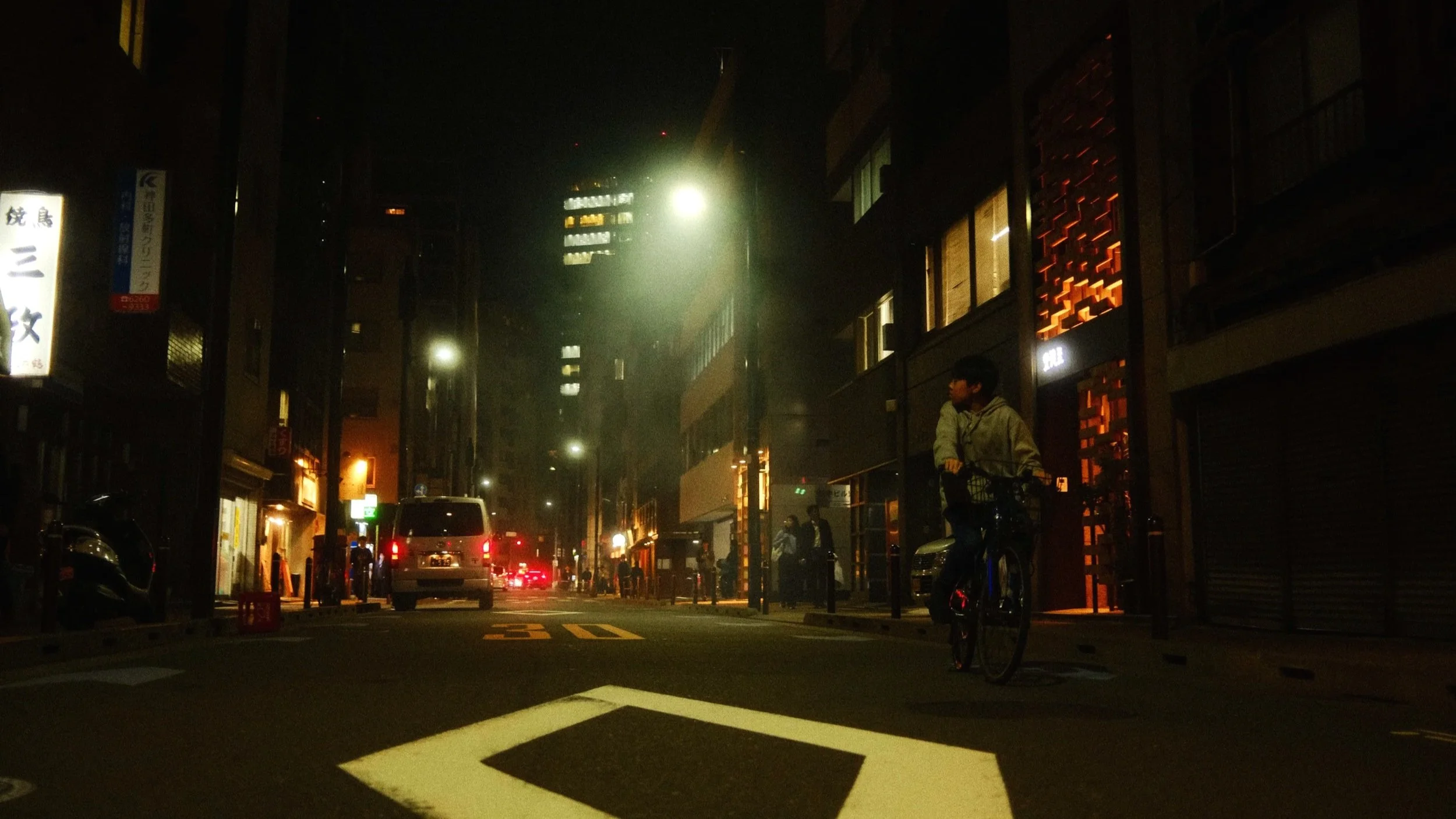 Nachtaufnahme einer Stadtstraße mit Autos, einem Radfahrer und Gebäuden mit leuchtenden Schildern und Fenstern.