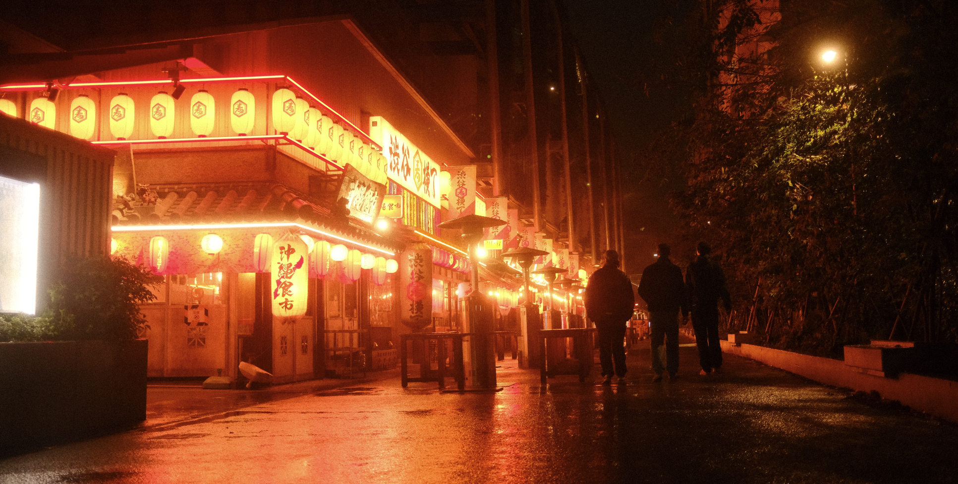 Drei Menschen gehen abends auf einer nassen, beleuchteten Straße an einem asiatisch inspirierten Restaurant mit roten Papierlaternen vorbei.