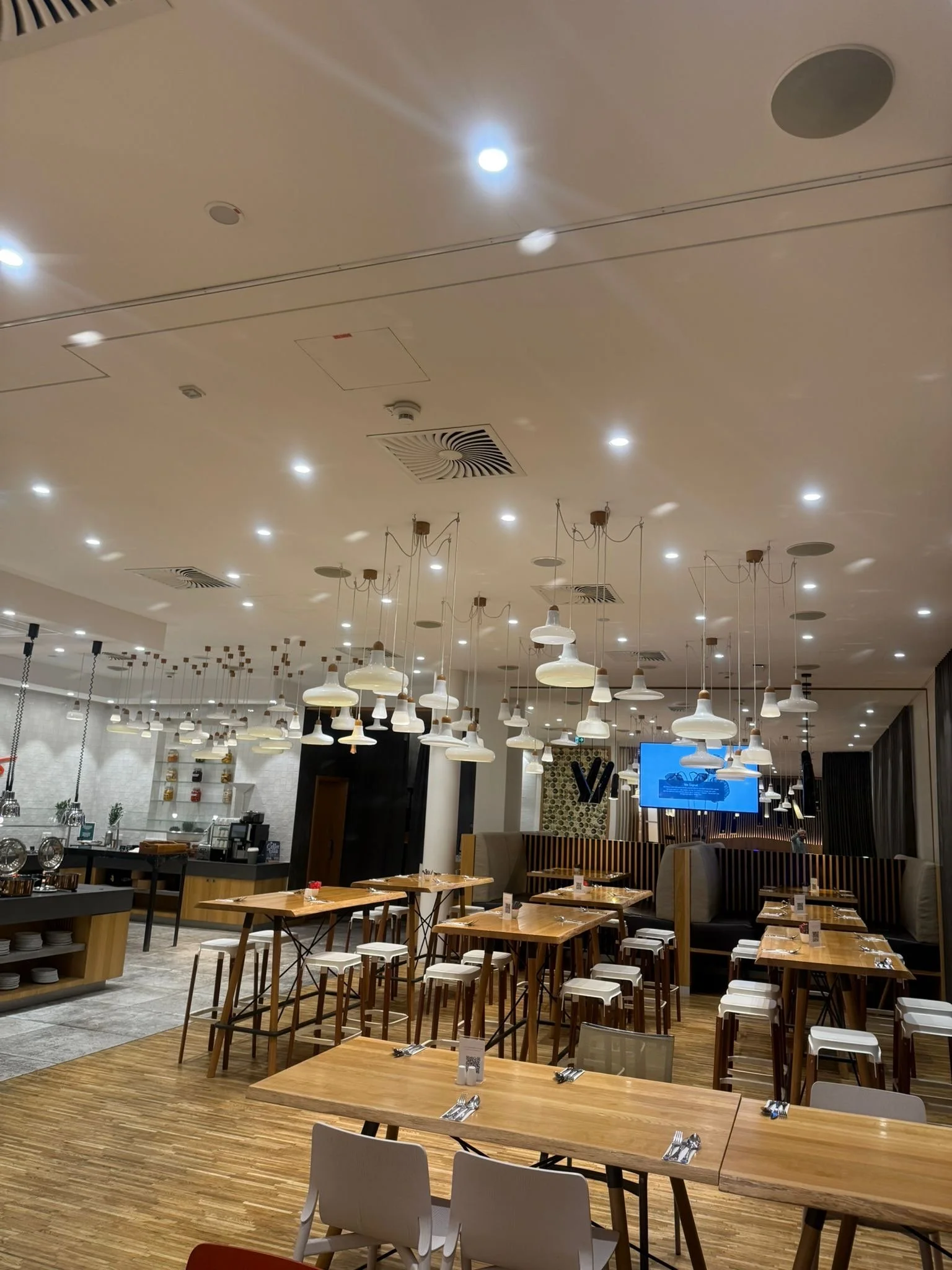 Interior of a modern restaurant with wooden tables and white chairs, pendant lights hanging from the ceiling, and a large television screen in the background.