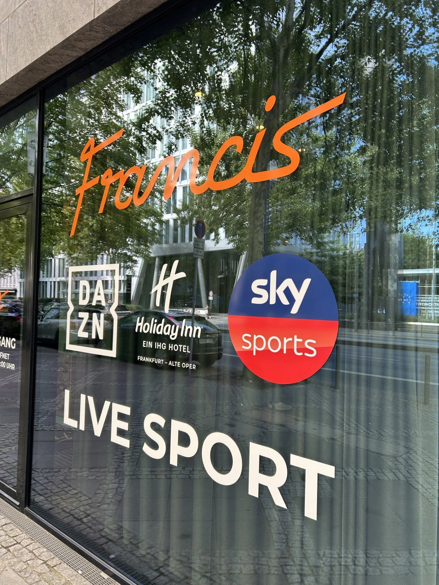 Store window displaying various logos and signs, including 'francis' in orange script, 'DAZN', 'Holiday Inn Frankfurt,' 'Sky Sports', and 'LIVE SPORT' in white letters.
